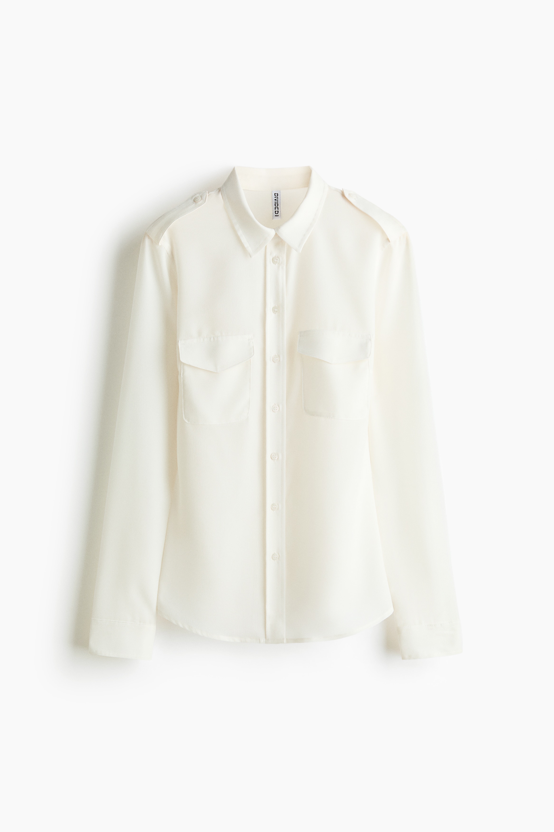 La modelo lleva una camisa tejida semitransparente en color blanco roto, con cuello, presillas abotonadas en los hombros y bolsillos de parche con solapa en el pecho. La camisa se lleva abierta sobre una camiseta grÃ¡fica blanca y una prenda de manga larga azul, y se combina con pantalones de chÃ¡ndal grises.