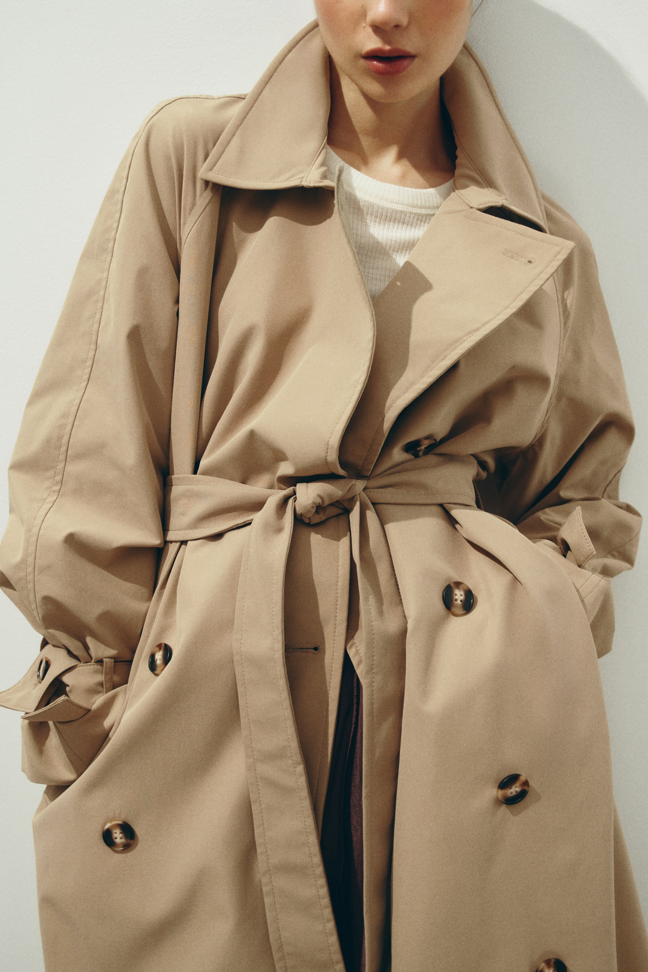 View larger image: A beige trench coat features wide, pointed lapels and a self-fabric belt tied at the waist, facing slightly forward. Tortoiseshell buttons detail the front panels, and large side pockets are visible, with a white ribbed top layered underneath.