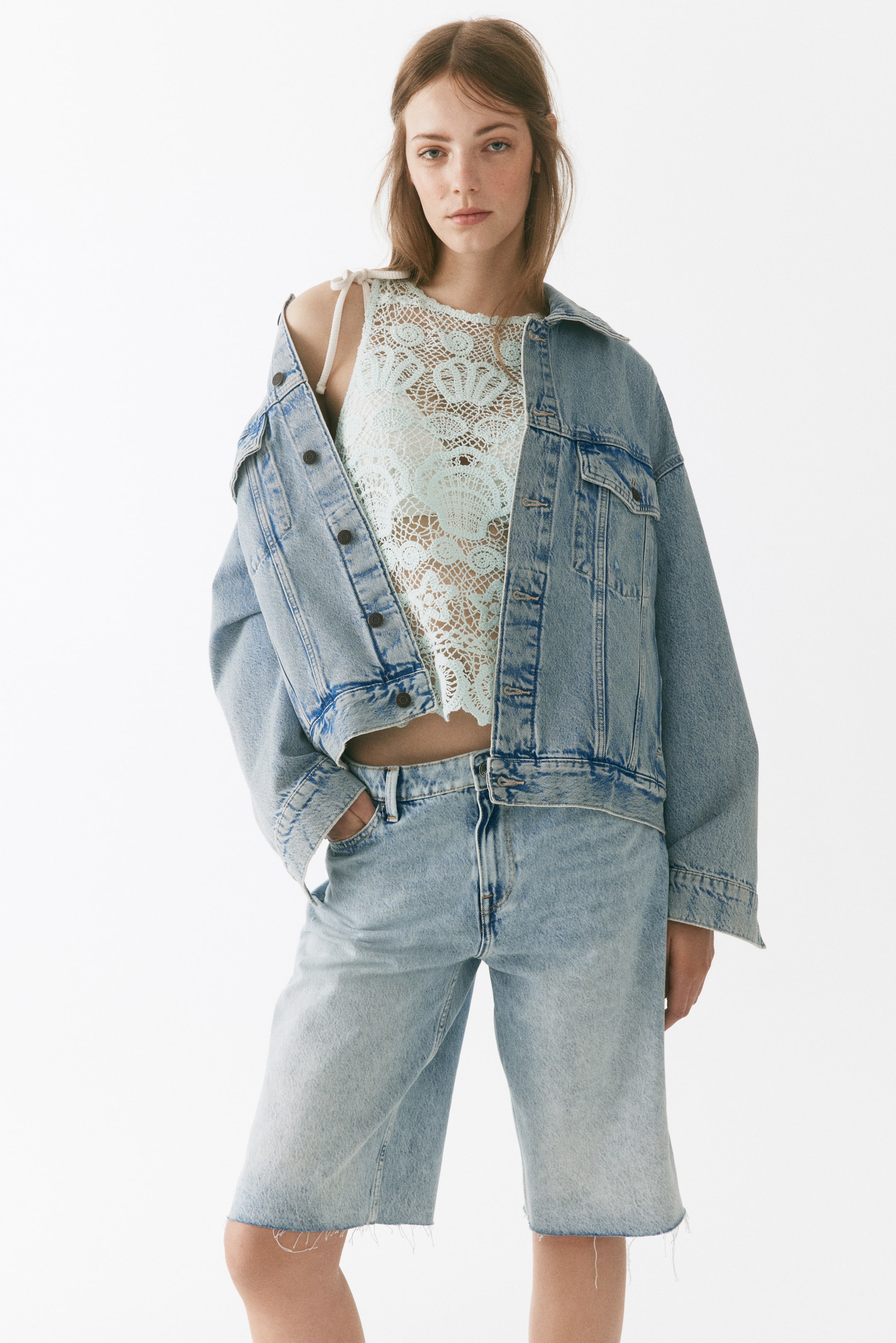 Visa större bild: En blek mintgrön virkad magtröja med ett intrikat öppet mönster med snäckliknande motiv. Den är designad med knytbara axelband och bärs under en öppen denimjacka, vilket framhäver dess korta passform.
