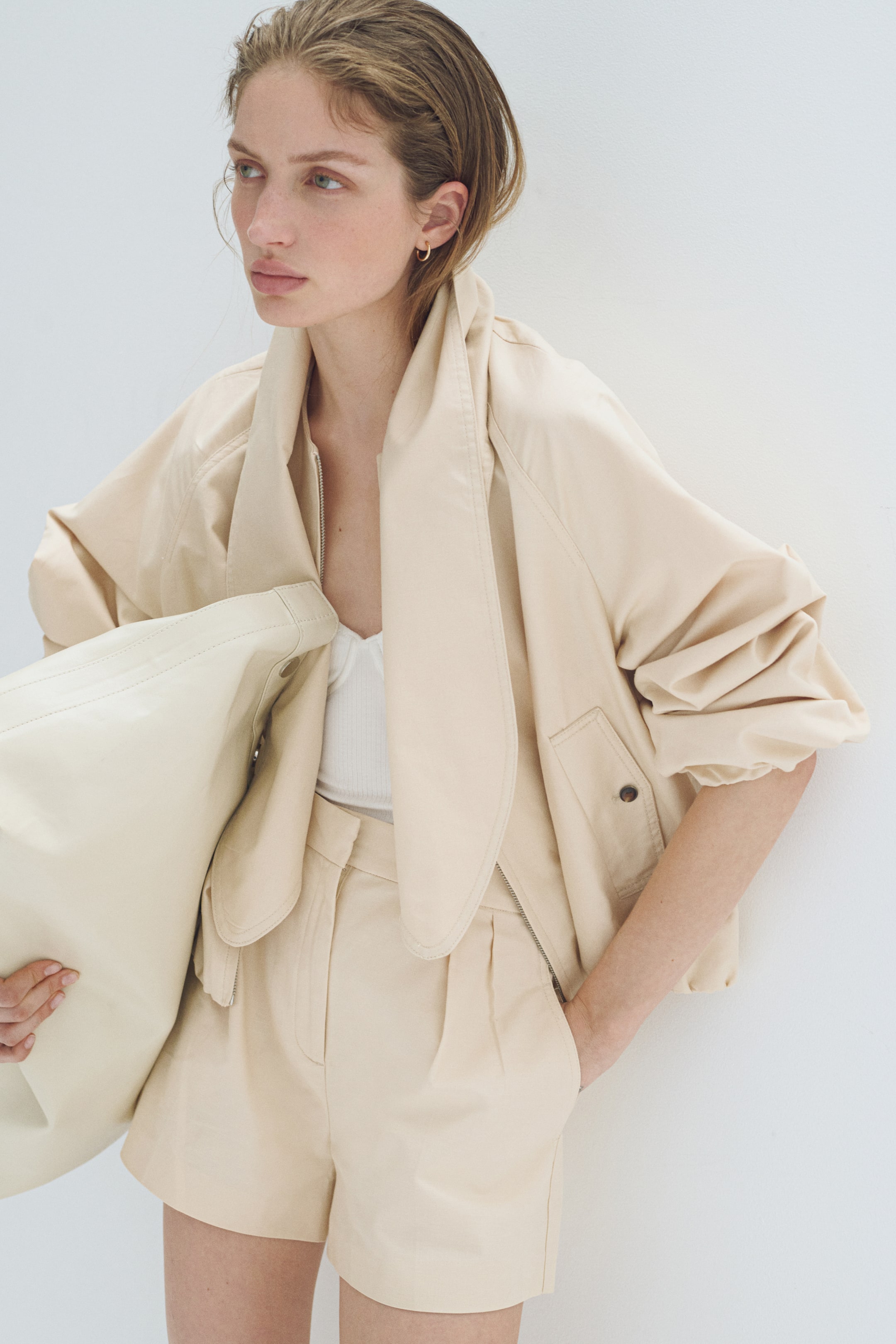 הצגת תמונה גדולה יותר: ז'קט קצר בצבע בז' בהיר מתאפיין בצווארון דרייפי גדול, סגירה קדמית חלקית עם רוכסן פתוח ושרוולים נפוחים, נלבש מעל חולצה לבנה עם טקסטורה מצולעת. מכנסיים קצרים תואמים בגזרה גבוהה עם קפלים משלימים את המראה, מציגים קפלים קדמיים וכיסי צד, בעוד תיק תואם בצבע בז' בהיר מוחזק כנגד הז'קט.