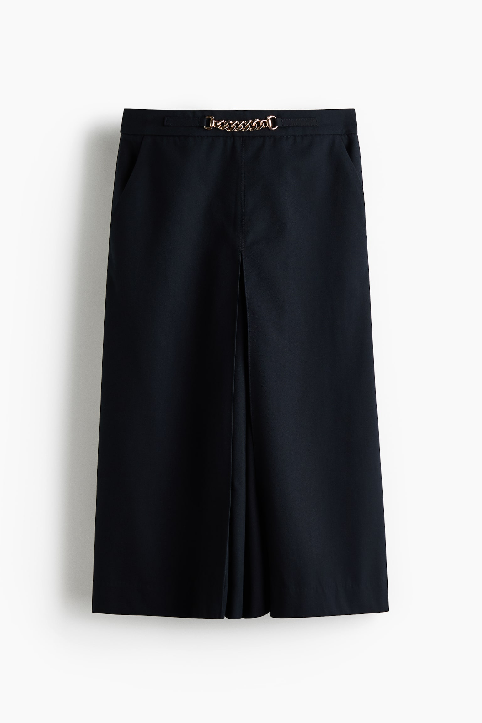 Koyu mavi midi boy culottes, ilmeklerden geçirilmiş altın tonlu zincir detayıyla düz bir bel bandı sergiliyor. Yan cepler kalçalarda görülebilir ve her geniş bacağın önünden aşağıya doğru keskin bir orta pli uzanıyor. - 1