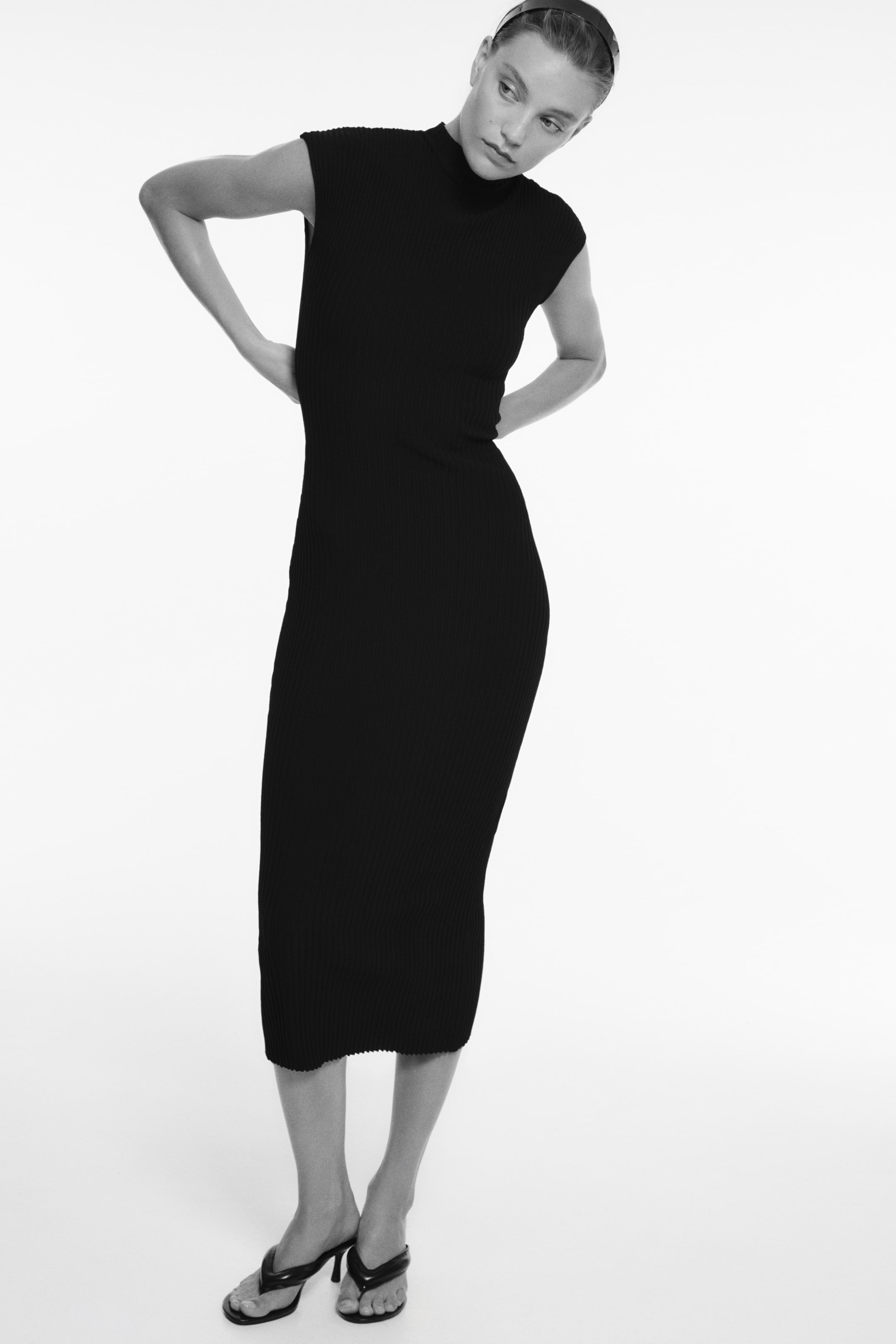 Ver imagen más grande: Una modelo lleva un vestido negro ajustado hasta media pantorrilla, confeccionado en canalé de punto de alta densidad, que realza su silueta ceñida al cuerpo desde el frente. El vestido presenta un cuello alto vuelto, manga corta con hombro ligeramente marcado y un bajo recto.