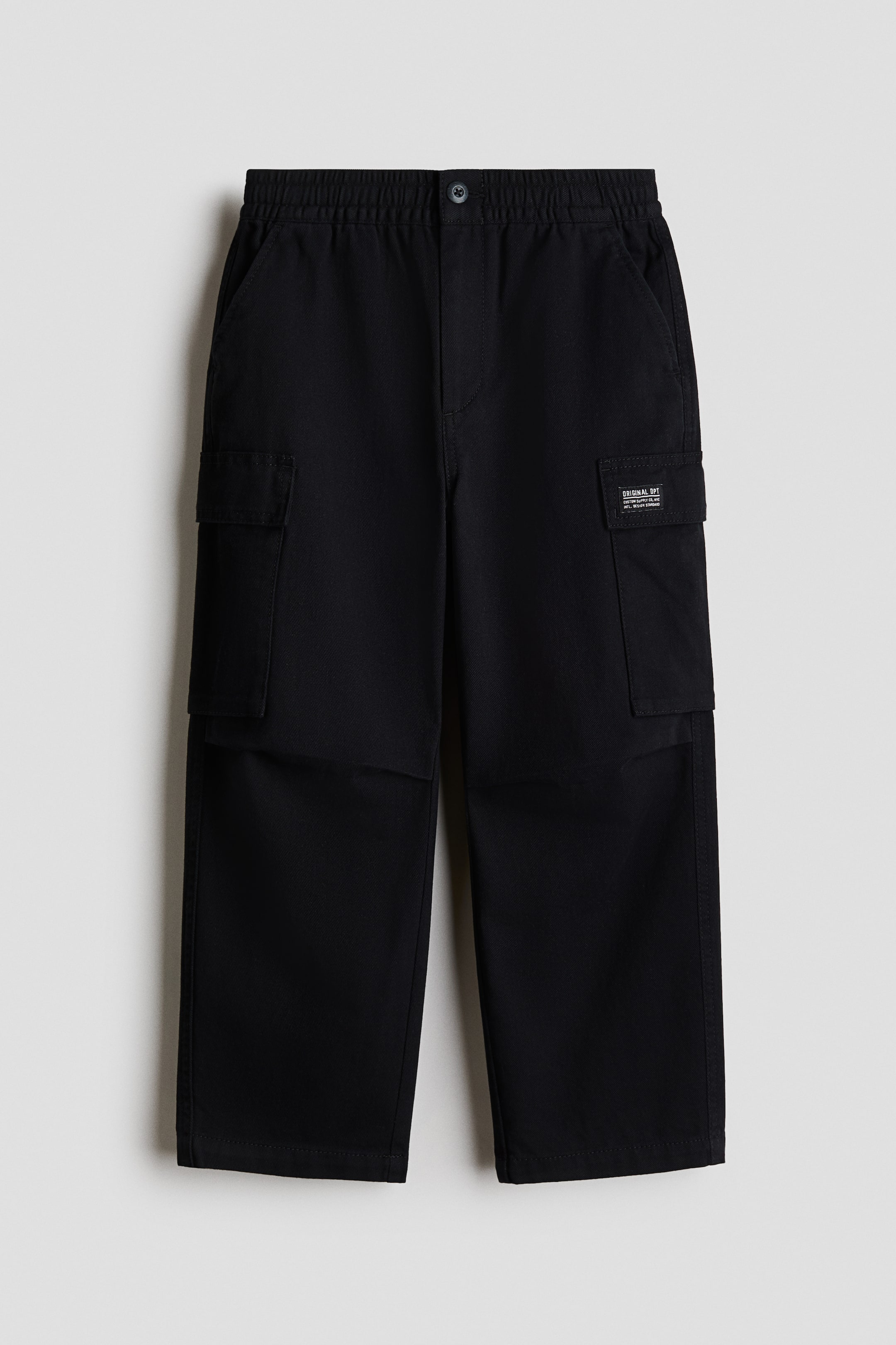 View larger image: Black cargo trousers are displayed from the front, featuring an elasticated waistband with a single button fastening. They are designed with two side slash pockets and two flap cargo pockets, the right cargo pocket showcasing a small black label.