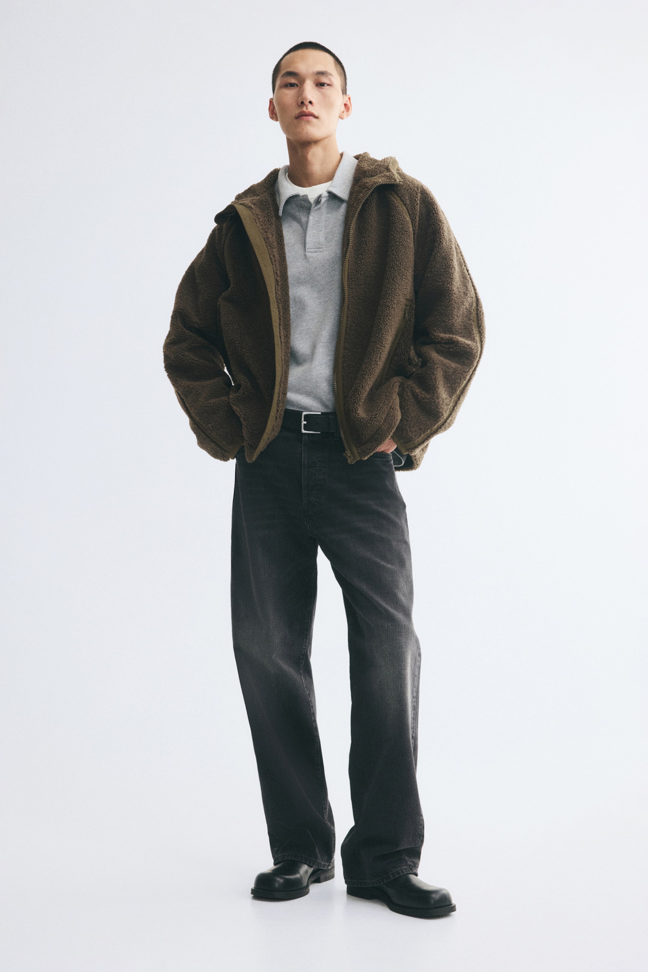 Agrandir l'image: Une veste en teddy fleece marron est portée ouverte par le mannequin, superposée sur un polo gris clair et un jean gris foncé. La veste présente une finition douce et texturée, une capuche et de longues manches raglan.