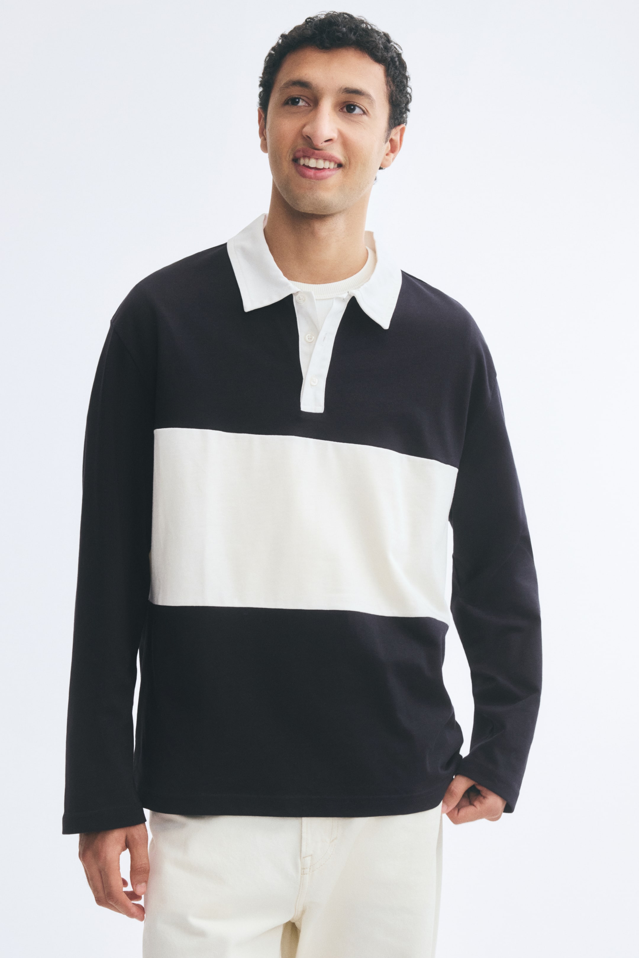 View larger image: A long-sleeved rugby shirt featuring a black body with a wide white horizontal stripe across the chest is being worn by the model. The shirt has a relaxed fit with dropped shoulders, a white collar, and a partially open button placket, styled over a white crew-neck t-shirt and paired with off-white trousers.