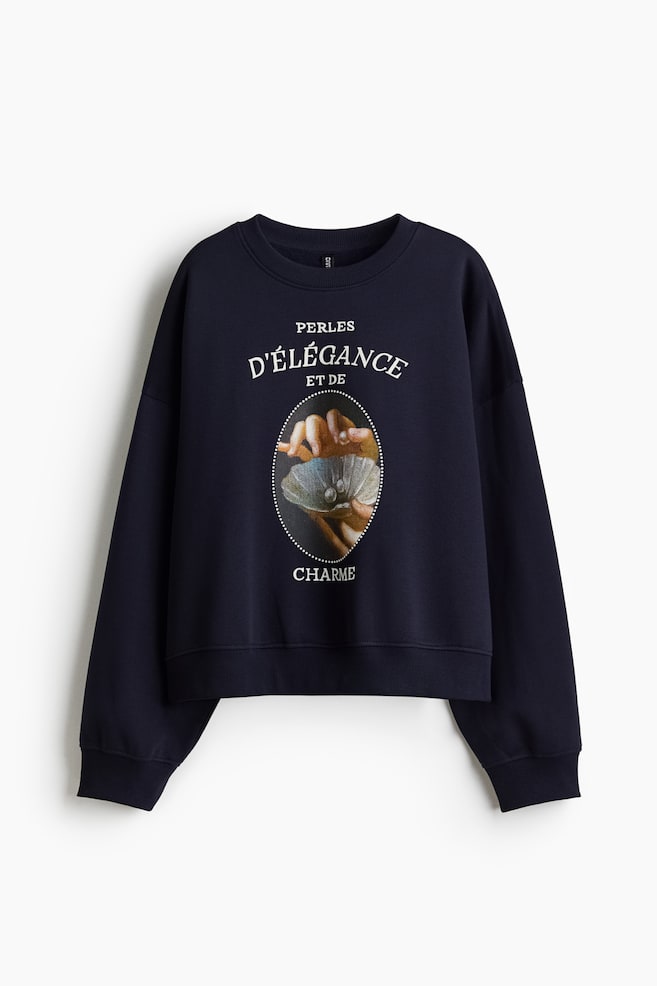 Un hanorac albastru marin este prezentat din FAȚĂ, având un decolteu rotund cu margine din nervuri, umeri căzuți, mâneci lungi cu manșete din nervuri și un tiv din nervuri. Un motiv central pe față afișează text alb și o imagine ovală cu mâini care țin o scoică deschisă cu perle. - 1