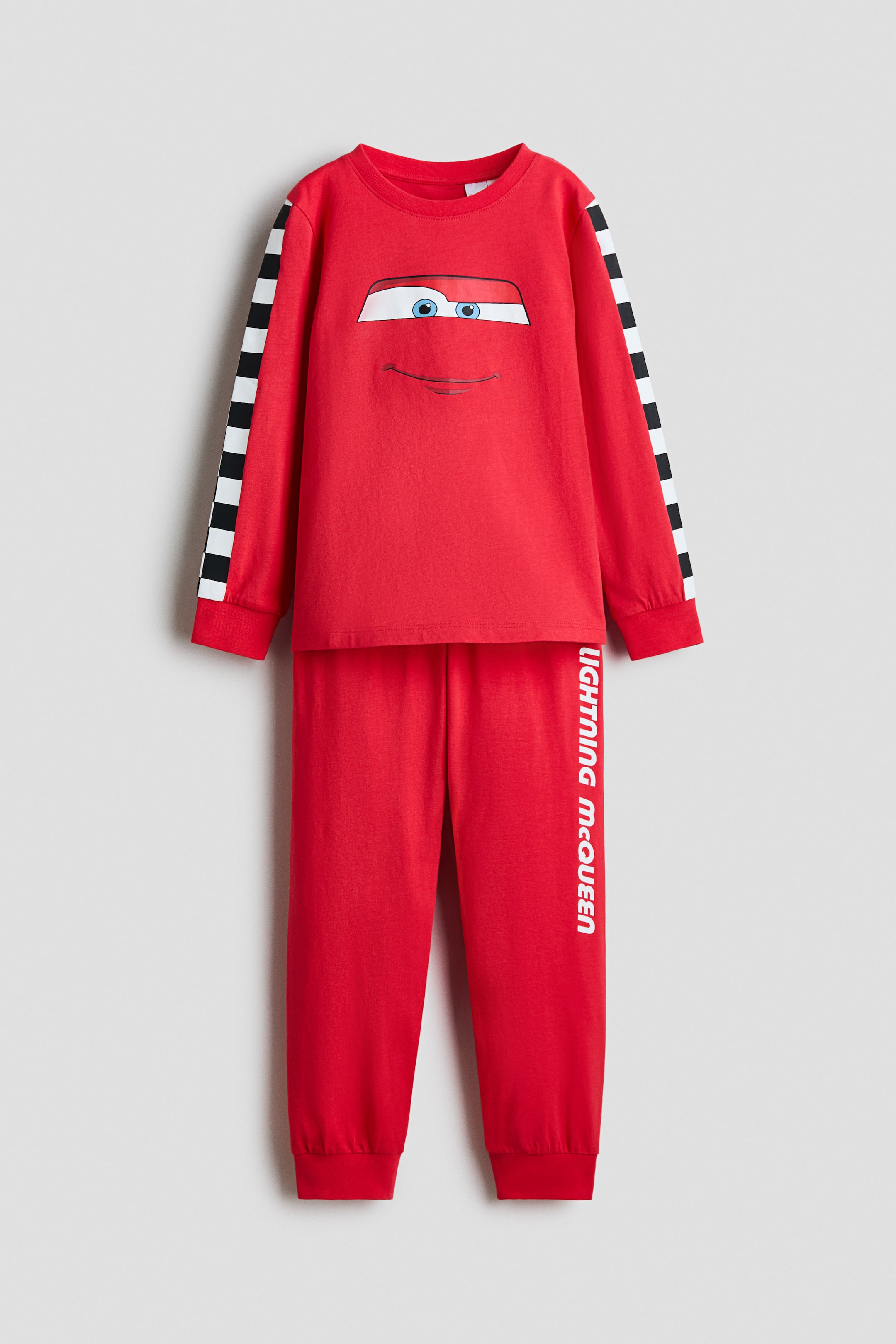 View larger image: A front-facing view shows a red cotton-jersey pajama set, with the long-sleeve top featuring a printed Lightning McQueen face design on the chest, black-and-white checkered stripes along the sleeves, and ribbed cuffs. The matching red pants display vertical white text reading "LIGHTNING McQUEEN" on the left leg and feature ribbed ankle cuffs.