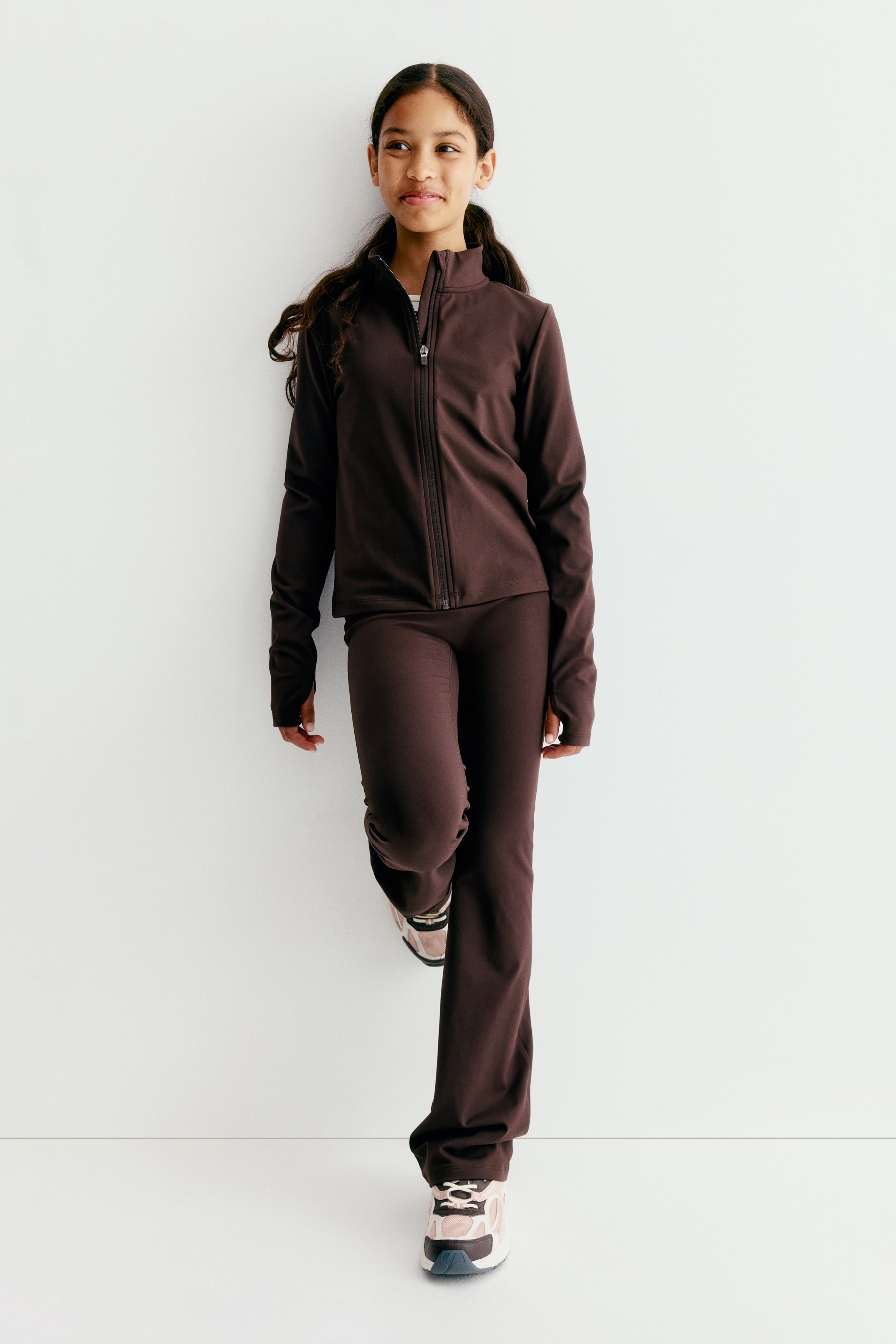 Une veste de sport marron foncé est présentée de face. Elle est dotée d'un col montant, d'une fermeture éclair intégrale à l'avant et de manches longues avec passe-pouces, et est coupée à une longueur raccourcie.