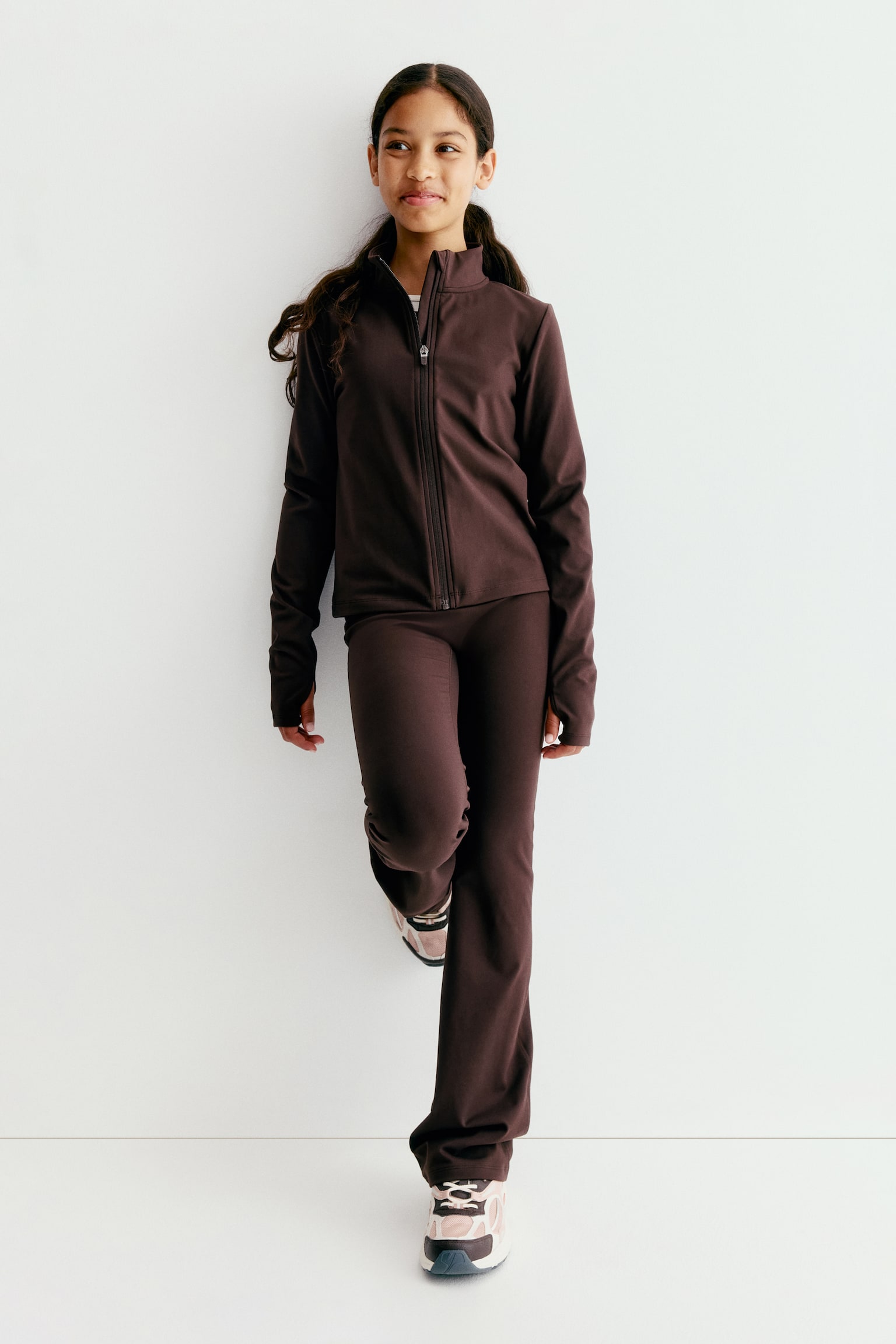 A dark brown sports jacket is showcasing a front-facing view. It boasts a mock neck, a full-zip front, and long sleeves with thumbholes, tailored to a cropped length. - 1