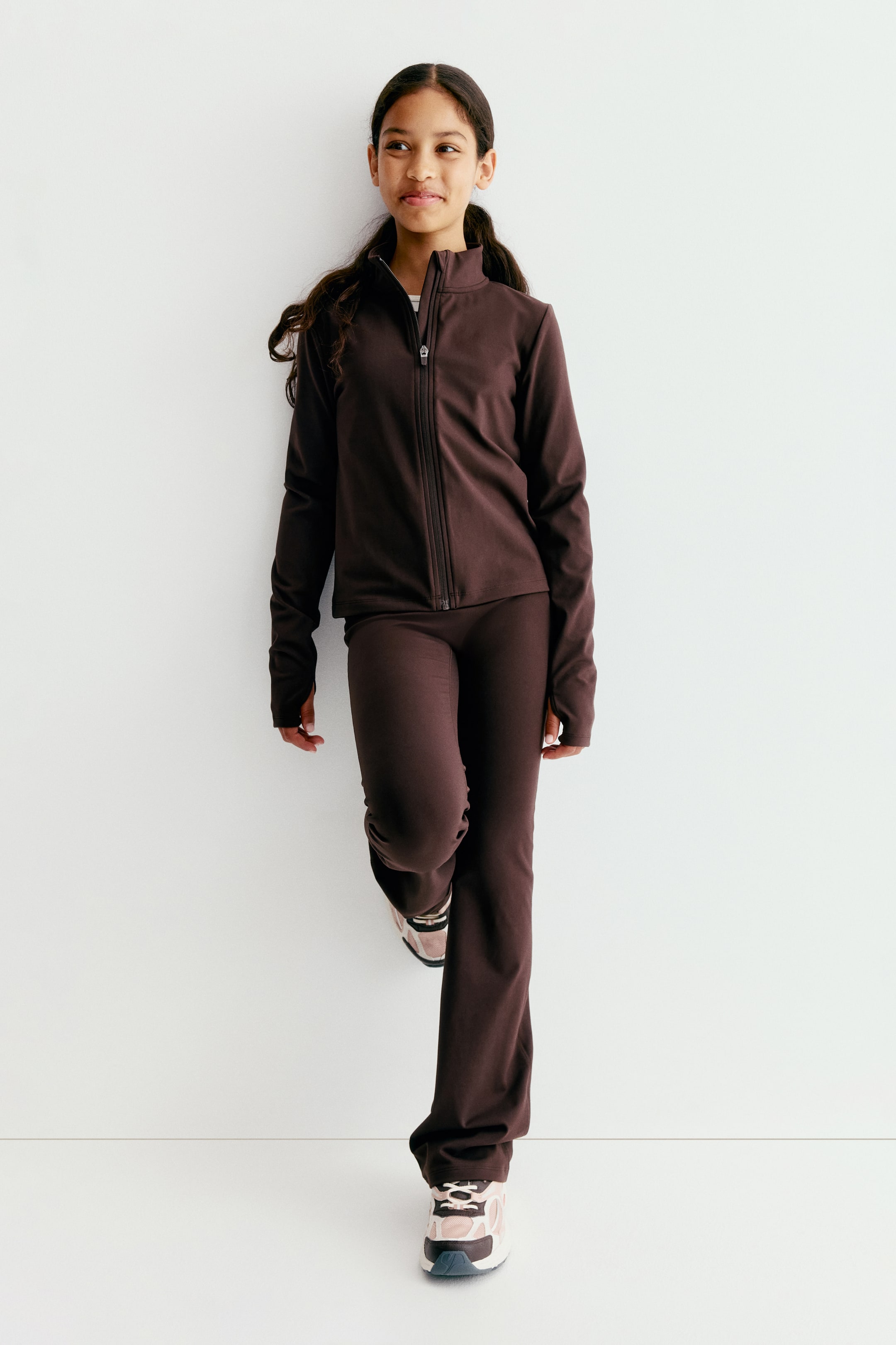 Grösseres Bild ansehen: Ein dunkelbraunes Activewear-Set wird von vorne präsentiert. Es besteht aus einer taillierten Jacke mit durchgehendem Reissverschluss, hohem Kragen und langen Ärmeln mit Daumenlochbündchen. Dazu werden passende, schmal geschnittene Hosen kombiniert, die am Saum leicht ausgestellt sind.