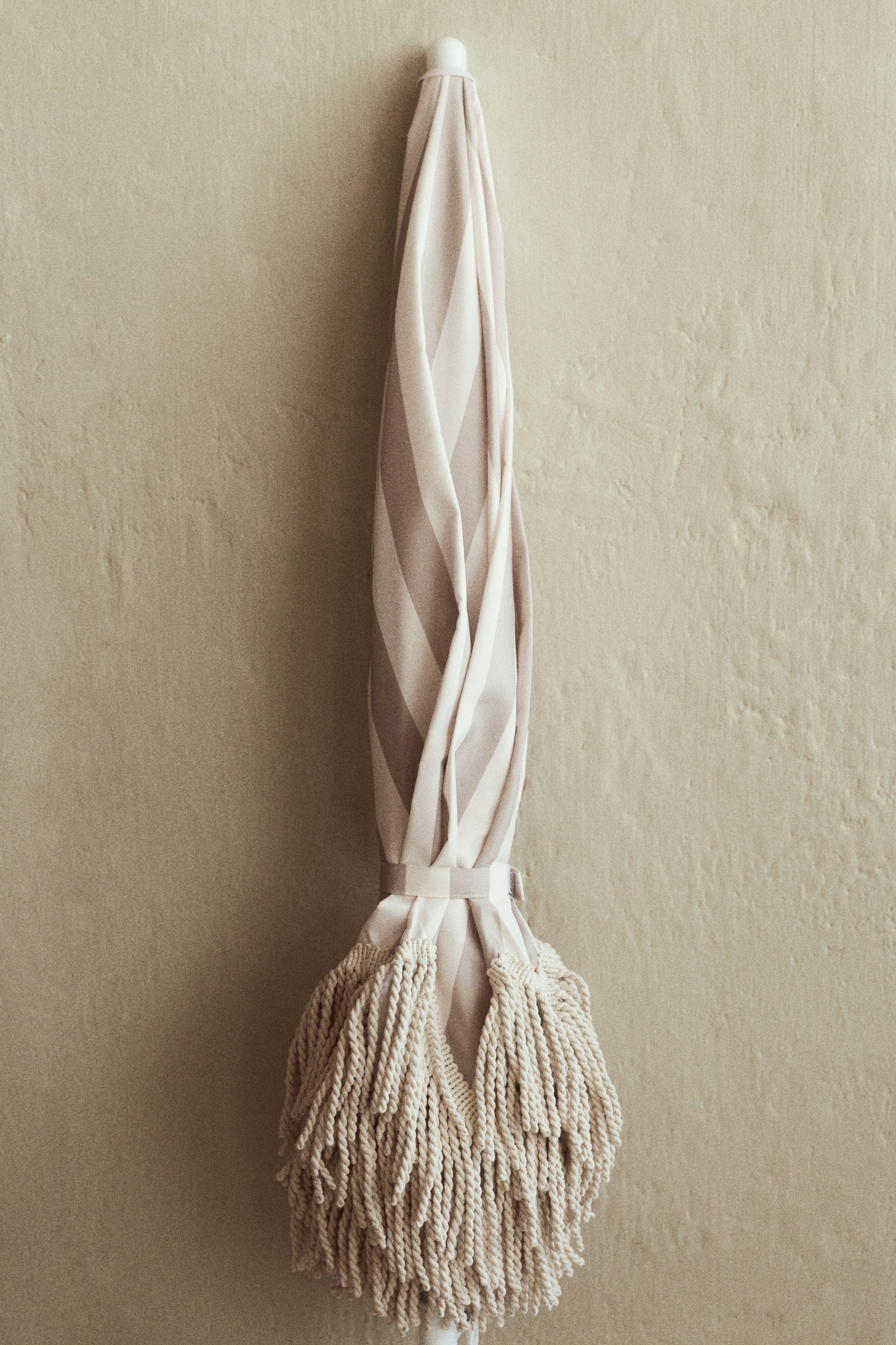 View larger image: A fringed parasol is presented vertically, folded and secured. Its fabric features alternating light and dark beige stripes, cinched by a hook-and-loop strap, with thick, braided fringes decorating the bottom portion. A white plastic pole is visible at its ends.