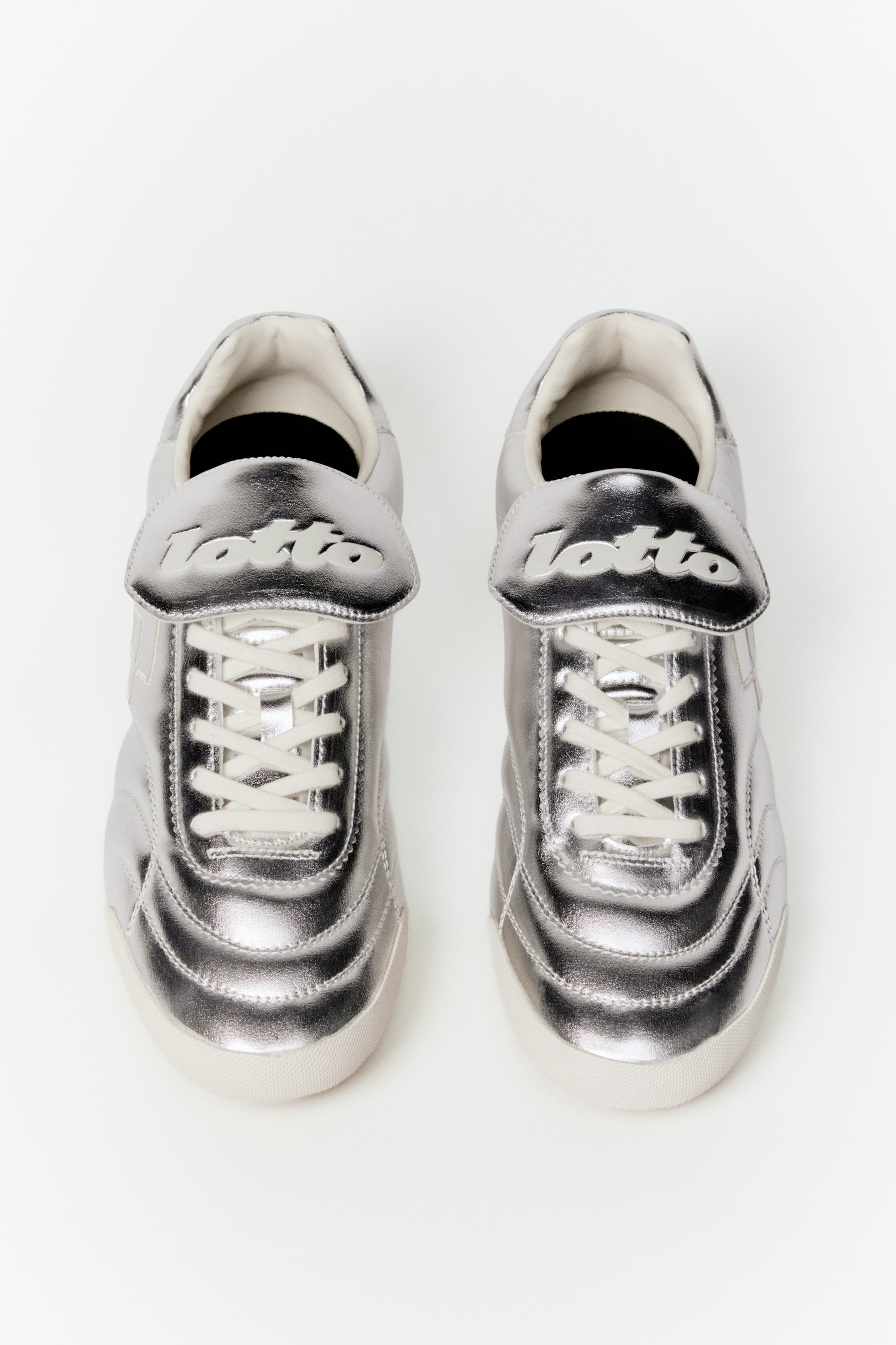View larger image: Two silver metallic sneakers are placed side-by-side, shown from an overhead perspective and facing forward. Each sneaker features white criss-cross laces beneath a large tongue flap embossed with the 'Lotto' logo, complemented by white soles and visible stitching detailing the upper panels.