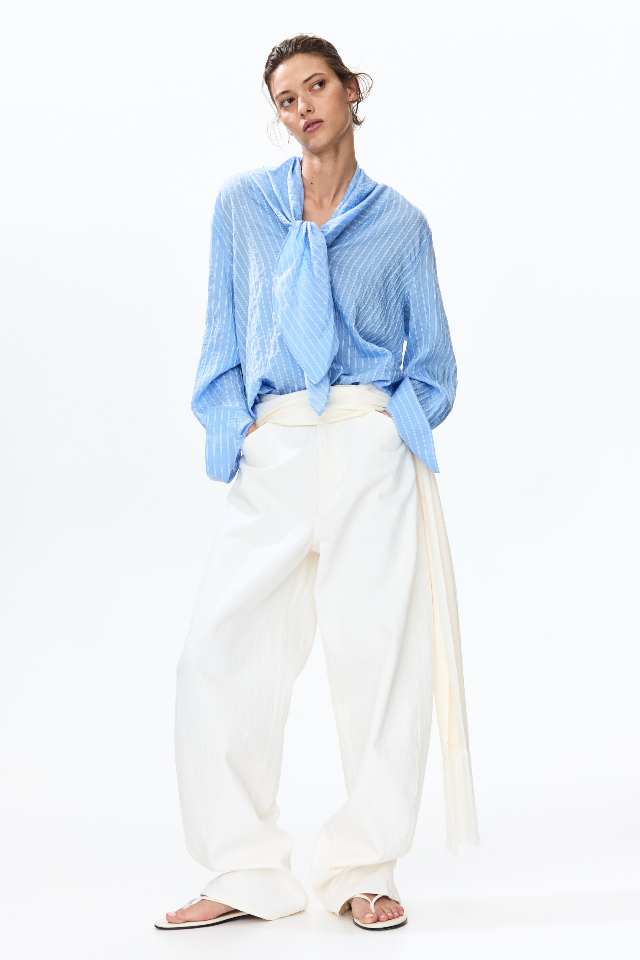 Agrandir l'image: Une blouse à rayures bleu clair avec une texture froissée et un grand nœud au cou est portée, rentrée dans un pantalon blanc à jambes larges avec une texture verticale subtile et une large ceinture enroulée formant une longue écharpe sur le côté droit. Des sandales blanches à lanières sont portées.