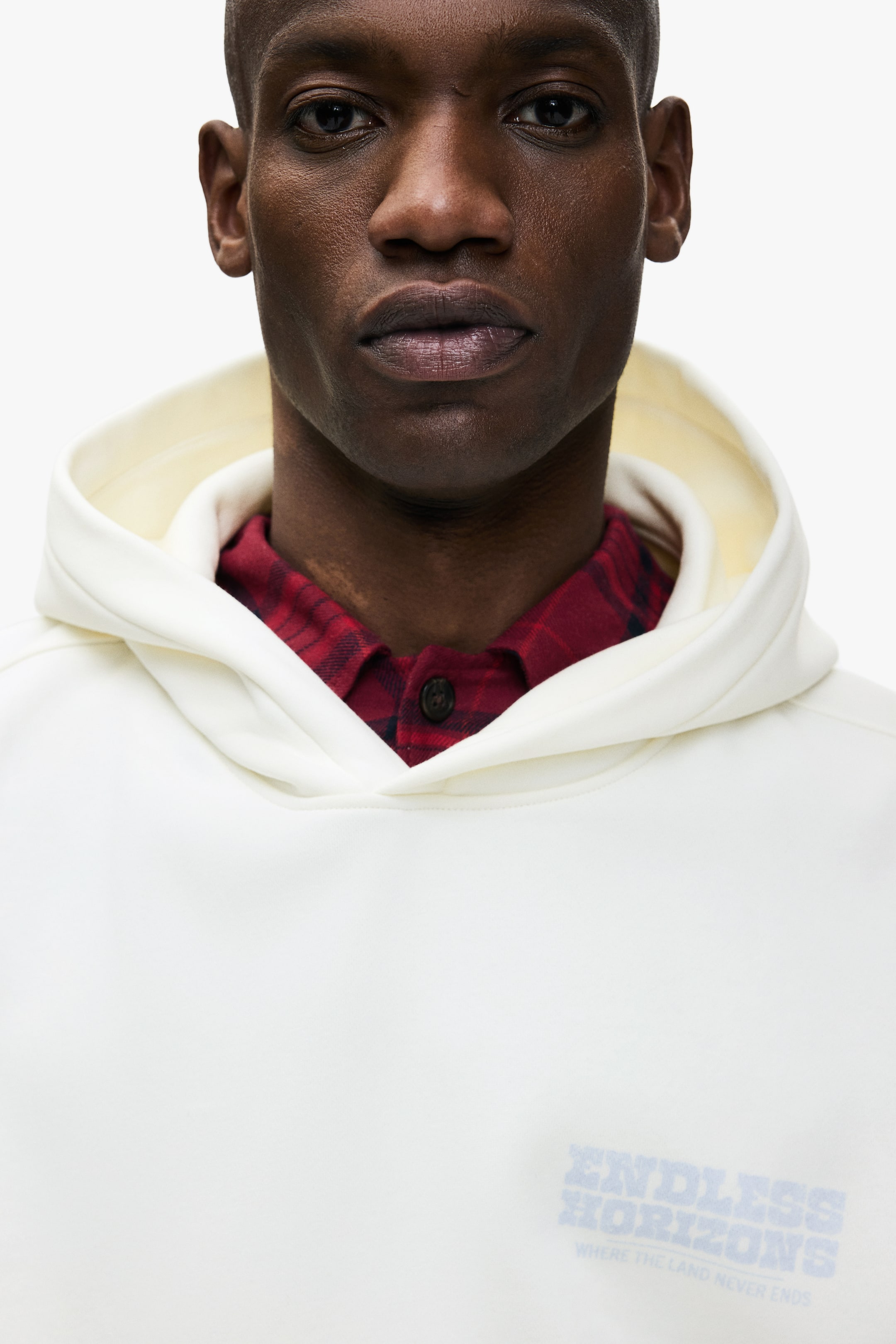 View larger image: A cream hoodie, featuring a visible hood and a faded blue graphic print reading 'ENDLESS HORIZONS WHERE THE LAND NEVER ENDS' on the chest, is being worn by the model. A red plaid shirt is being layered beneath the hoodie, with its collar visible at the neckline.