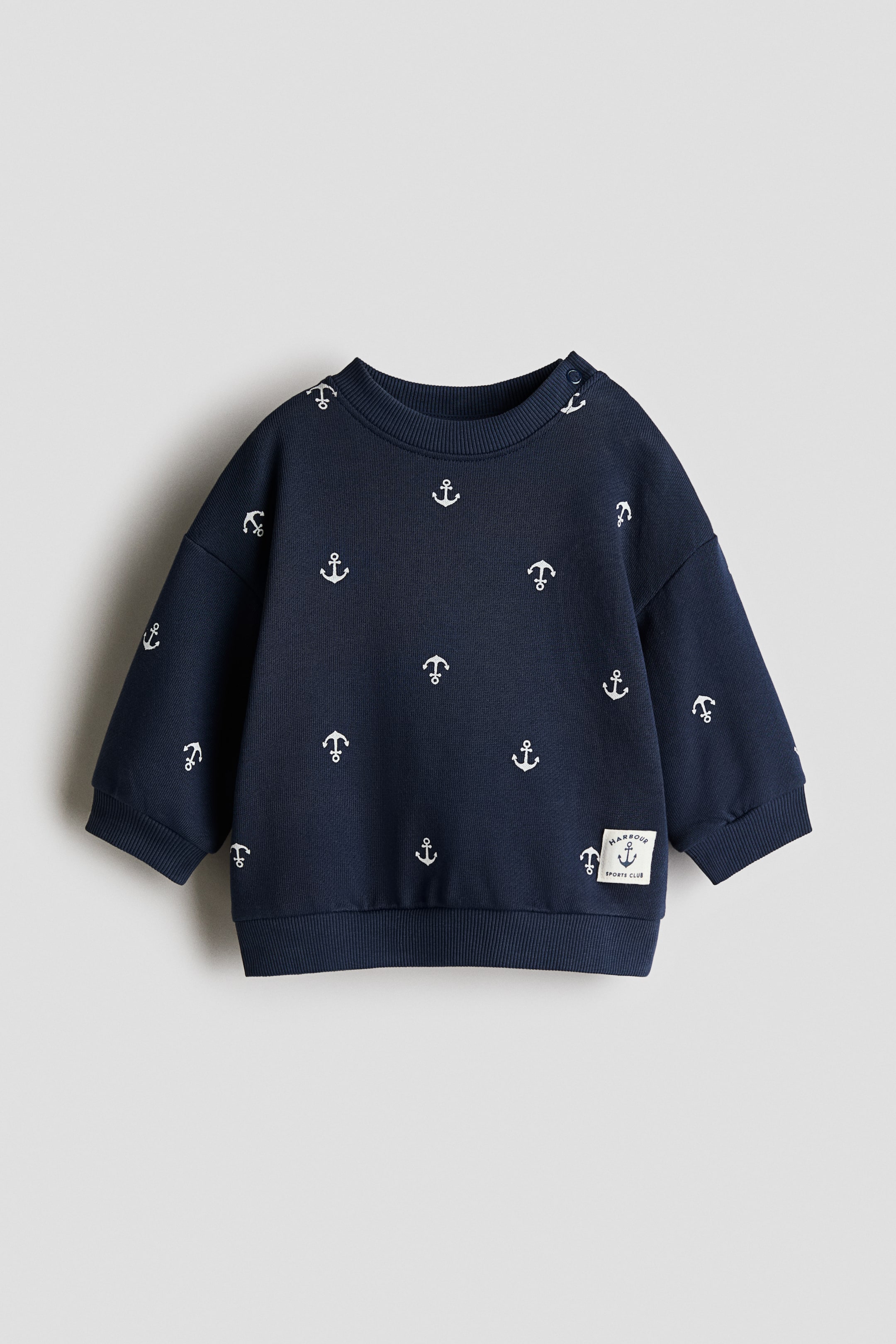 Grotere afbeelding bekijken: Een marineblauw sweatshirt met lange mouwen toont de voorkant en heeft een all-over patroon van witte ankermotieven. Het is uitgevoerd met een geribde ronde hals, drukknoopsluitingen op de linkerschouder, geribde manchetten en een geribde boord, met een "Harbour Sports Club"-patch linksonder.