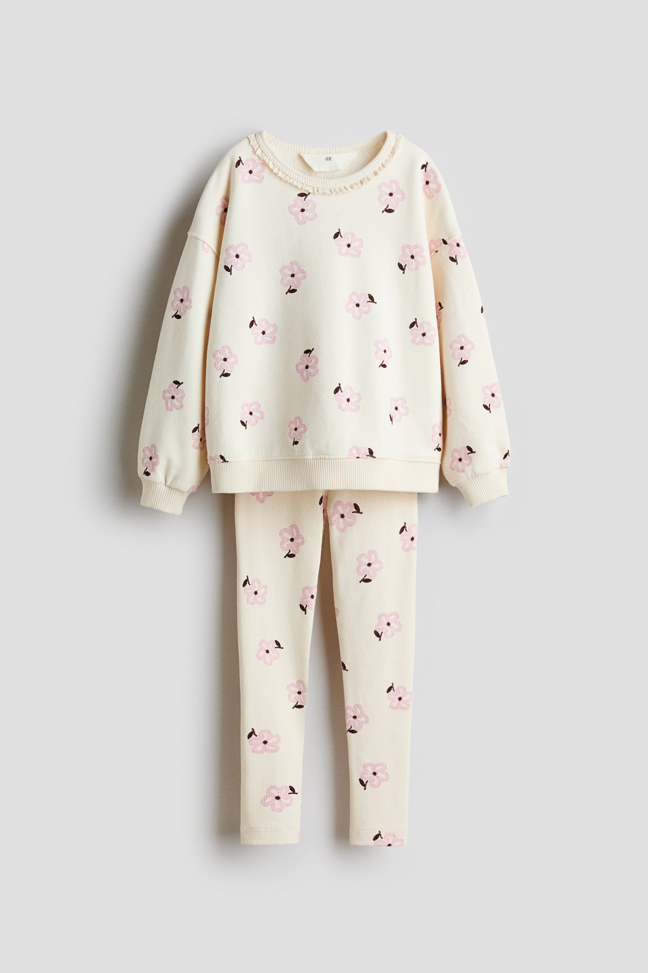 Ver imagen más grande: Un conjunto de dos piezas de frente compuesto por una sudadera color crema y leggings a juego de largo completo, ambos con un patrón repetido de flores rosas con centros oscuros. La sudadera presenta un ribete con volantes a lo largo de su cuello redondo, puños acanalados y un dobladillo acanalado.