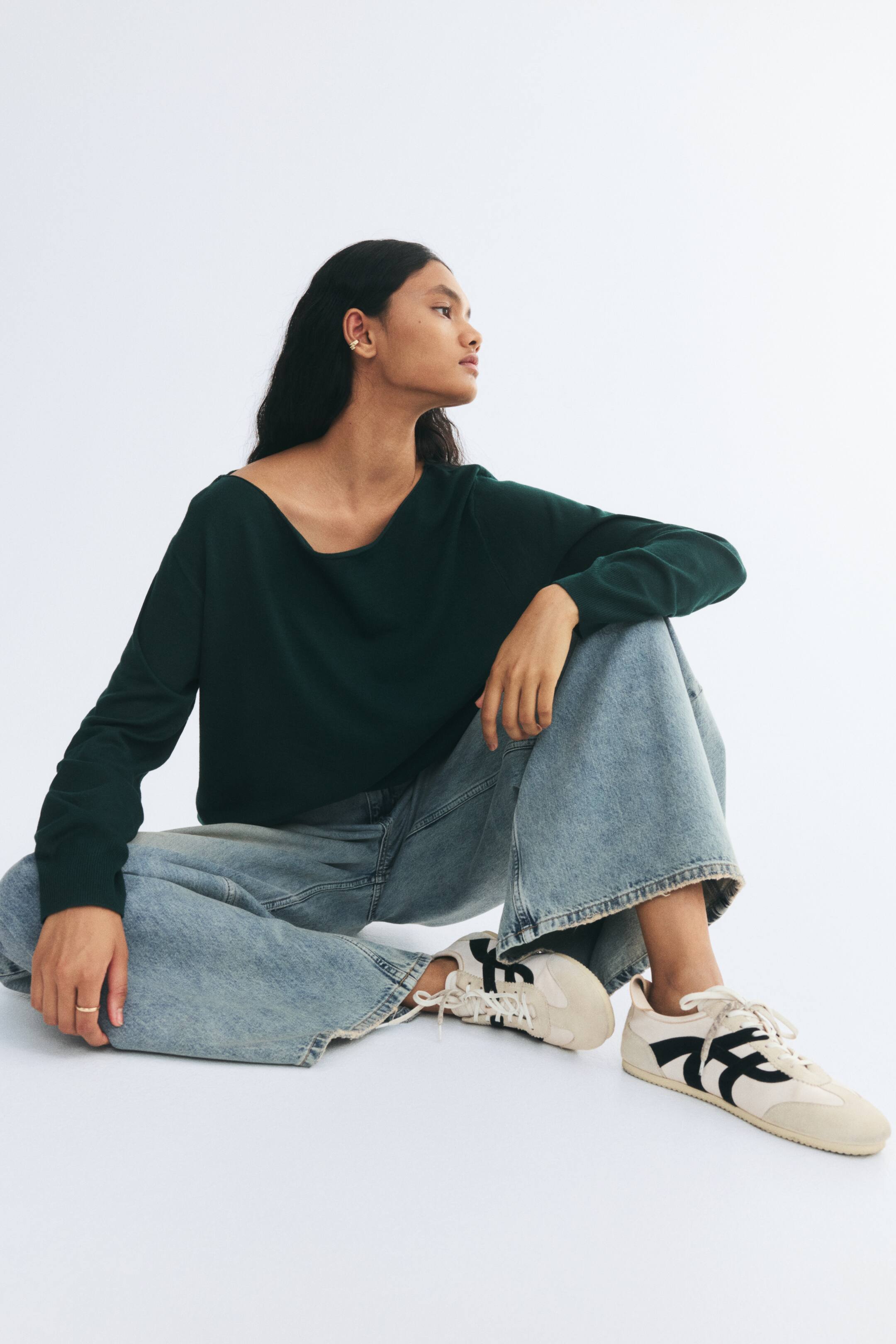 View larger image: A dark green, off-the-shoulder knit jumper featuring a rolled-edge neckline, dropped shoulders, and ribbed cuffs is being worn by the model. The jumper is styled with light-wash, wide-leg denim jeans and cream sneakers.