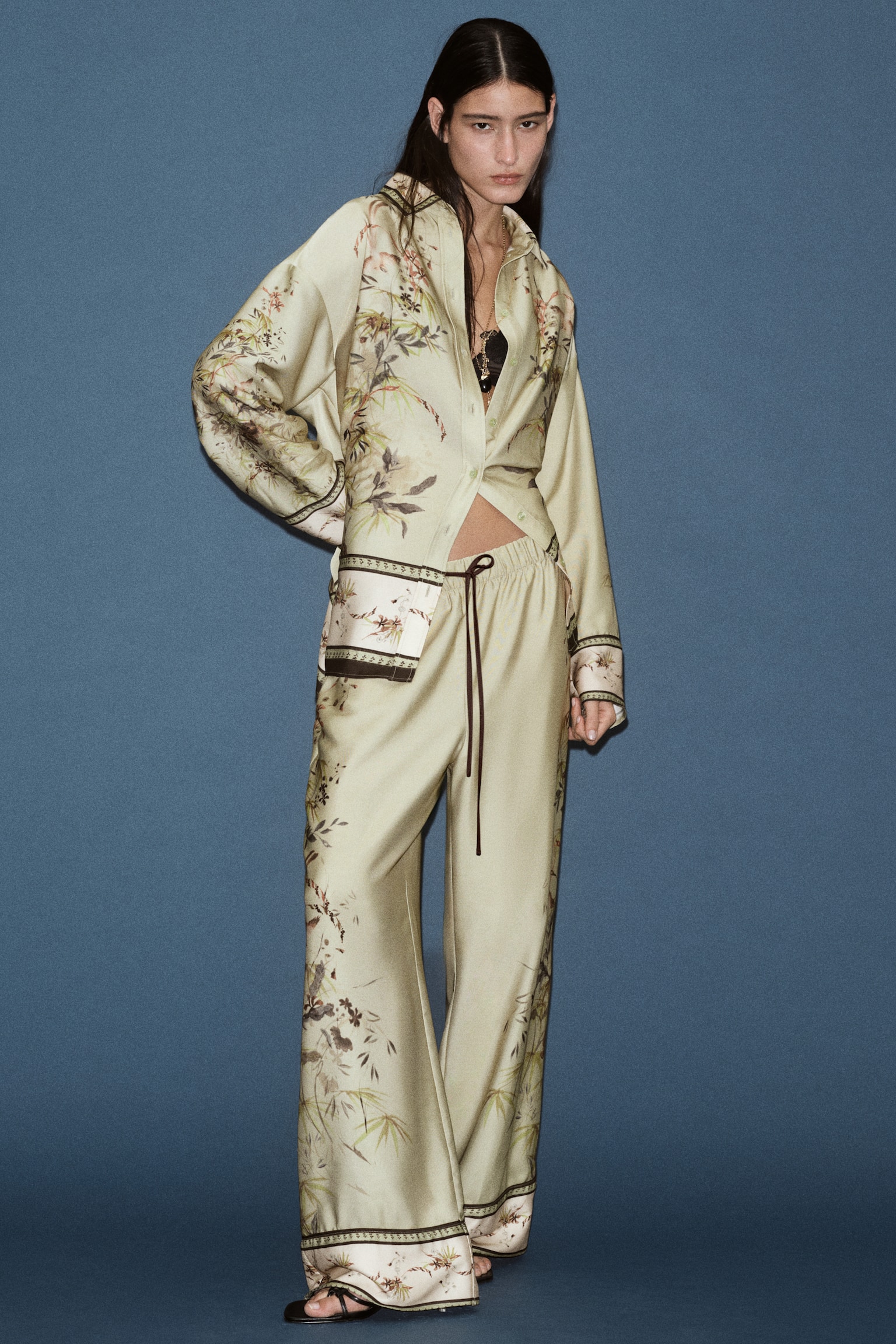 Trousers in a light green patterned weave are shown from the front, featuring an elasticated waist with a dark brown drawstring and wide, straight legs. The trousers display a nature-inspired print of birds and foliage cascading down the outer sides, finishing with a cream-coloured, double-layered border with dark piping trim at the hems. - 1