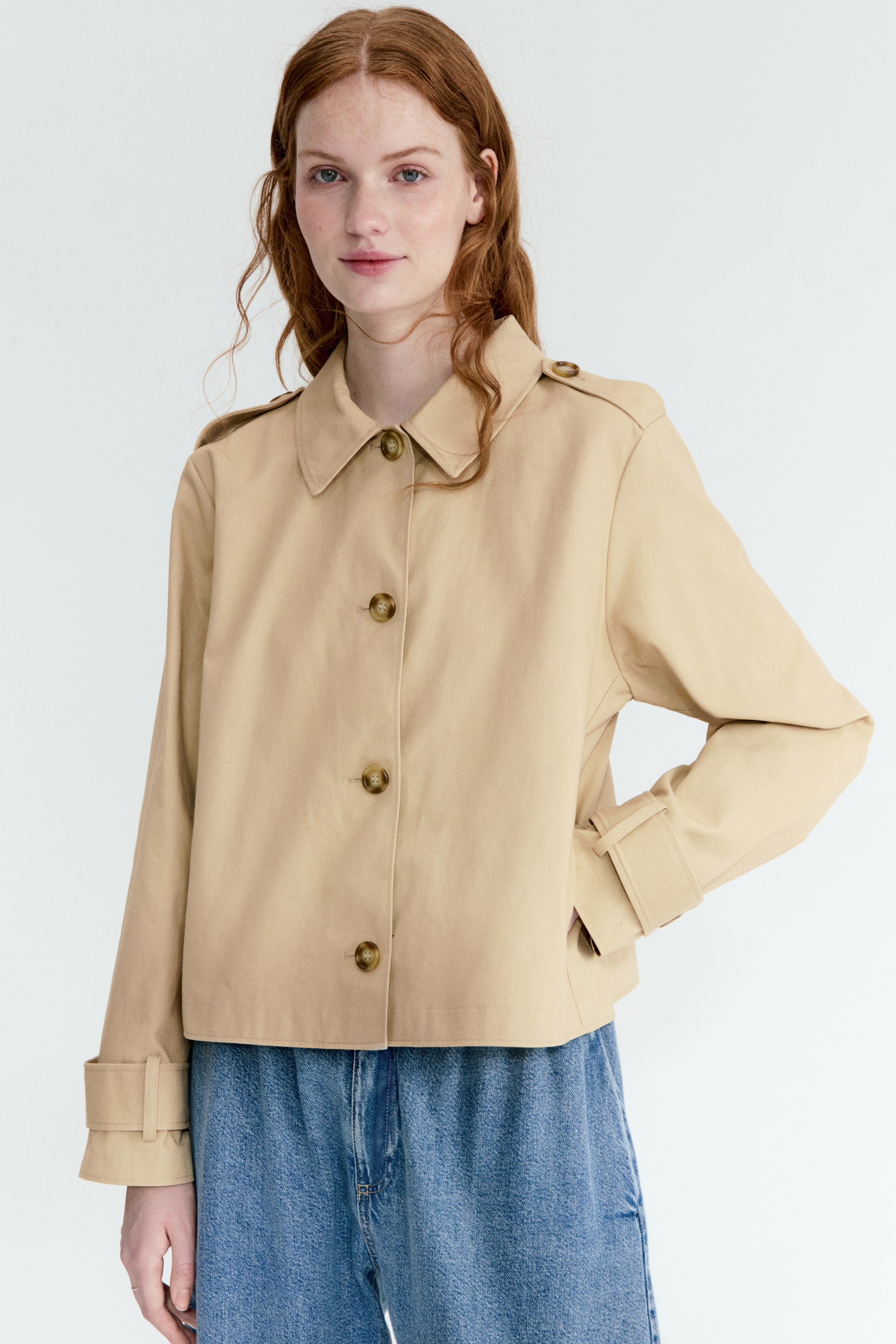 View larger image: A short beige jacket showcases a button-down front adorned with four brown buttons, a pointed collar, and epaulets on the shoulders. It features long sleeves with adjustable cuff straps, with one cuff rolled up, and visible side pockets.