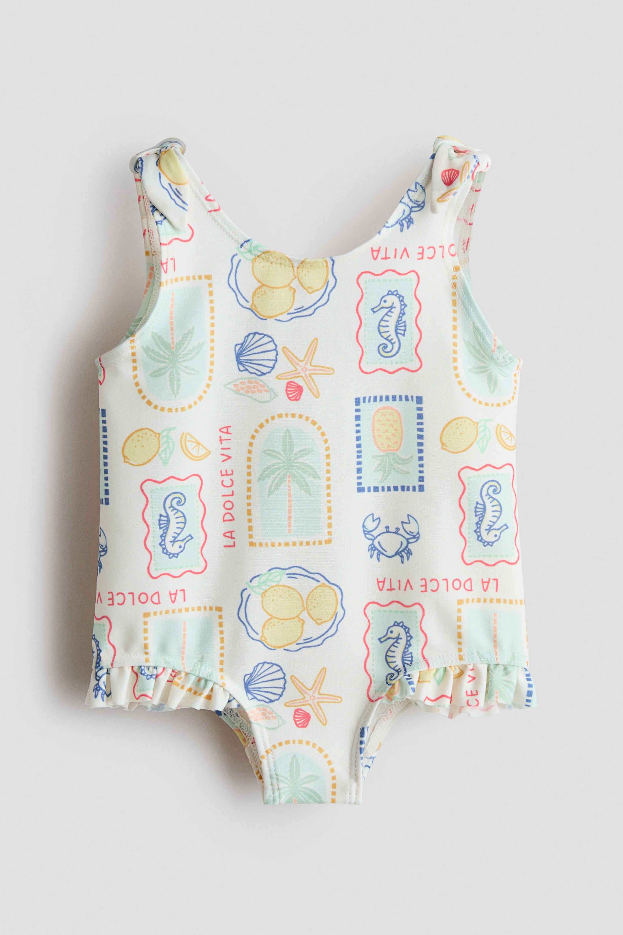 Agrandir l'image: Un maillot de bain une pièce blanc est présenté de face, arborant un motif répétitif de citrons pastel, de palmiers, d'hippocampes, de crabes, de coquillages et d'étoiles de mer, accompagné du texte "LA DOLCE VITA" dans des designs encadrés. Il est conçu avec des bretelles froncées et une bordure à volants ornant les ouvertures des jambes.