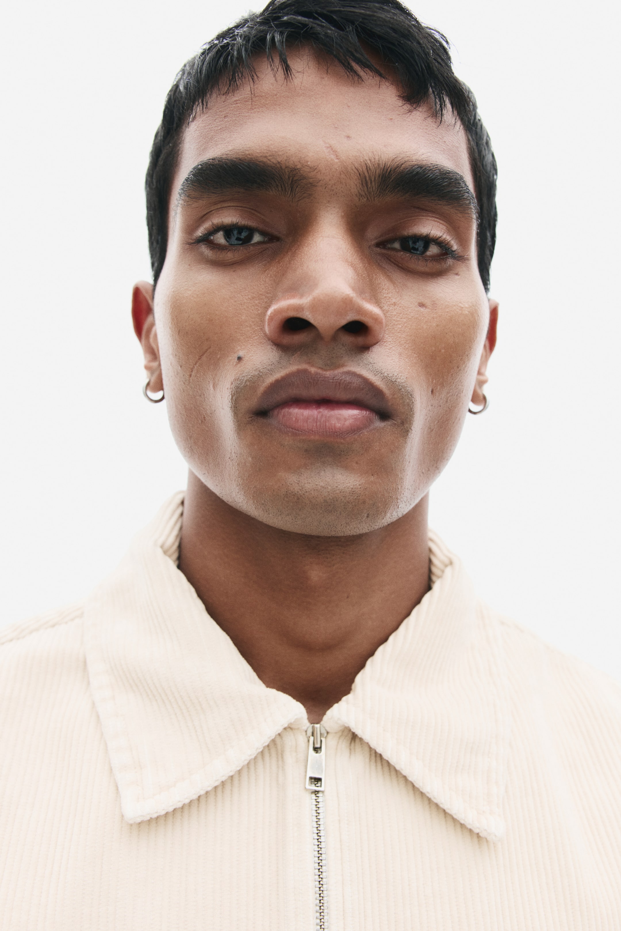 View larger image: The model is wearing a cream-coloured corduroy overshirt, shown in a close-up view. The shirt features a classic collar and a metallic front zip closure.