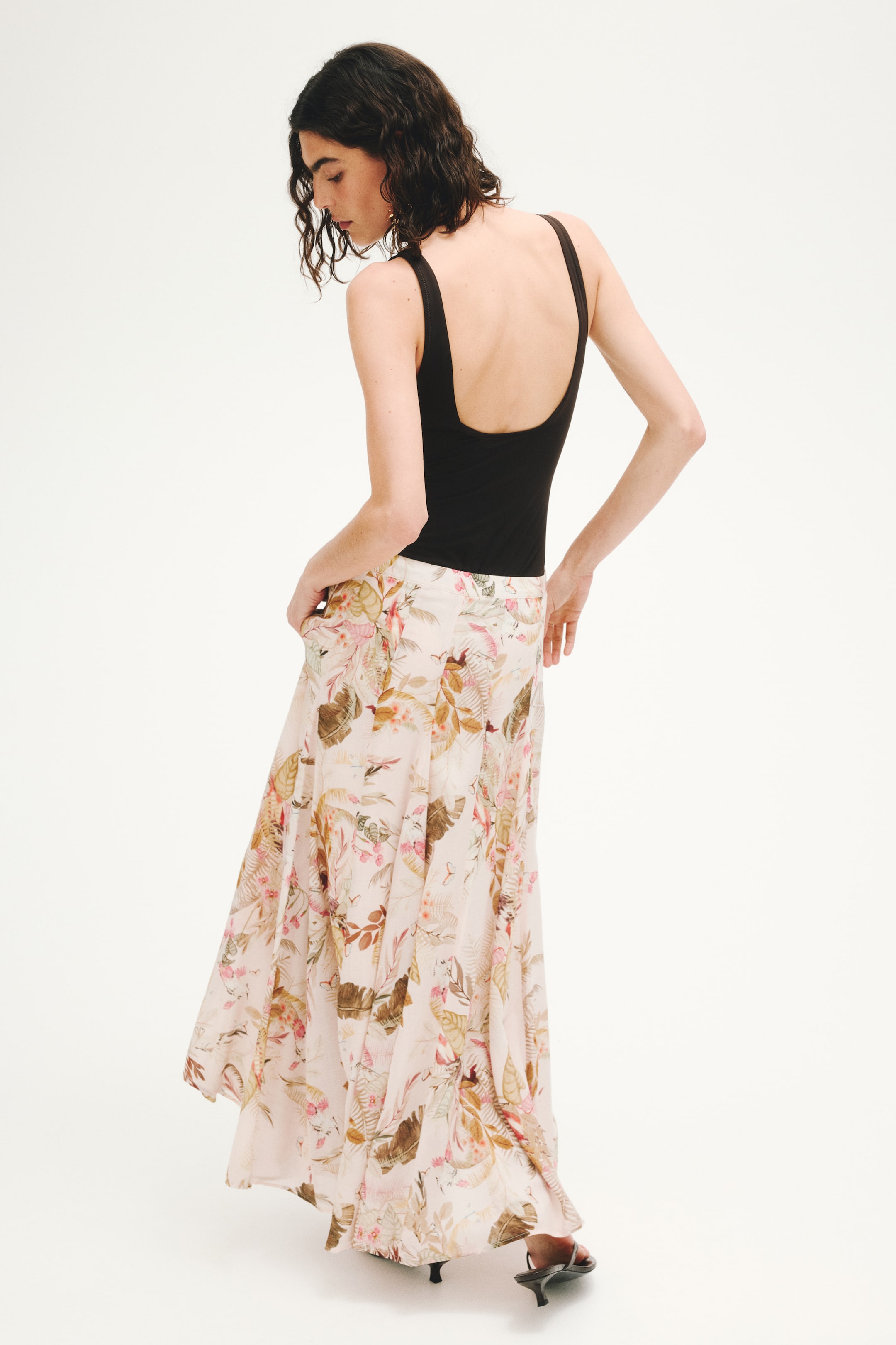 View larger image: A light pink maxi skirt, adorned with a tropical floral pattern in shades of brown, green, and pink, gracefully flows from a high waist. The skirt is displayed from a back-right angle, highlighting its complete silhouette and how it is styled with a tucked-in black top and black kitten heel sandals.