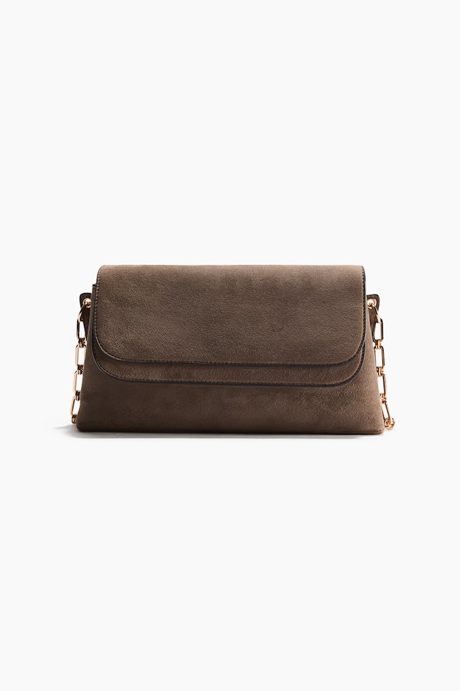A brown suede-textured shoulder bag is facing front, showcasing a rectangular silhouette and a layered top flap with dark-trimmed edges. Gold-toned metal chain straps are connecting to the sides of the bag. - 1