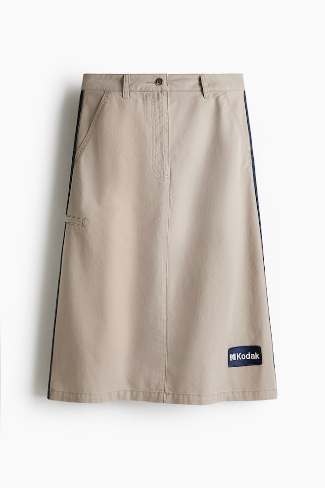 A beige midi skirt is hanging, featuring a flat waistband with belt loops, a single button closure, and two angled front pockets. It is detailed with a dark contrast stripe running down each side seam, a small horizontal loop on the left side, and a rectangular Kodak brand patch on the lower right front. - 1