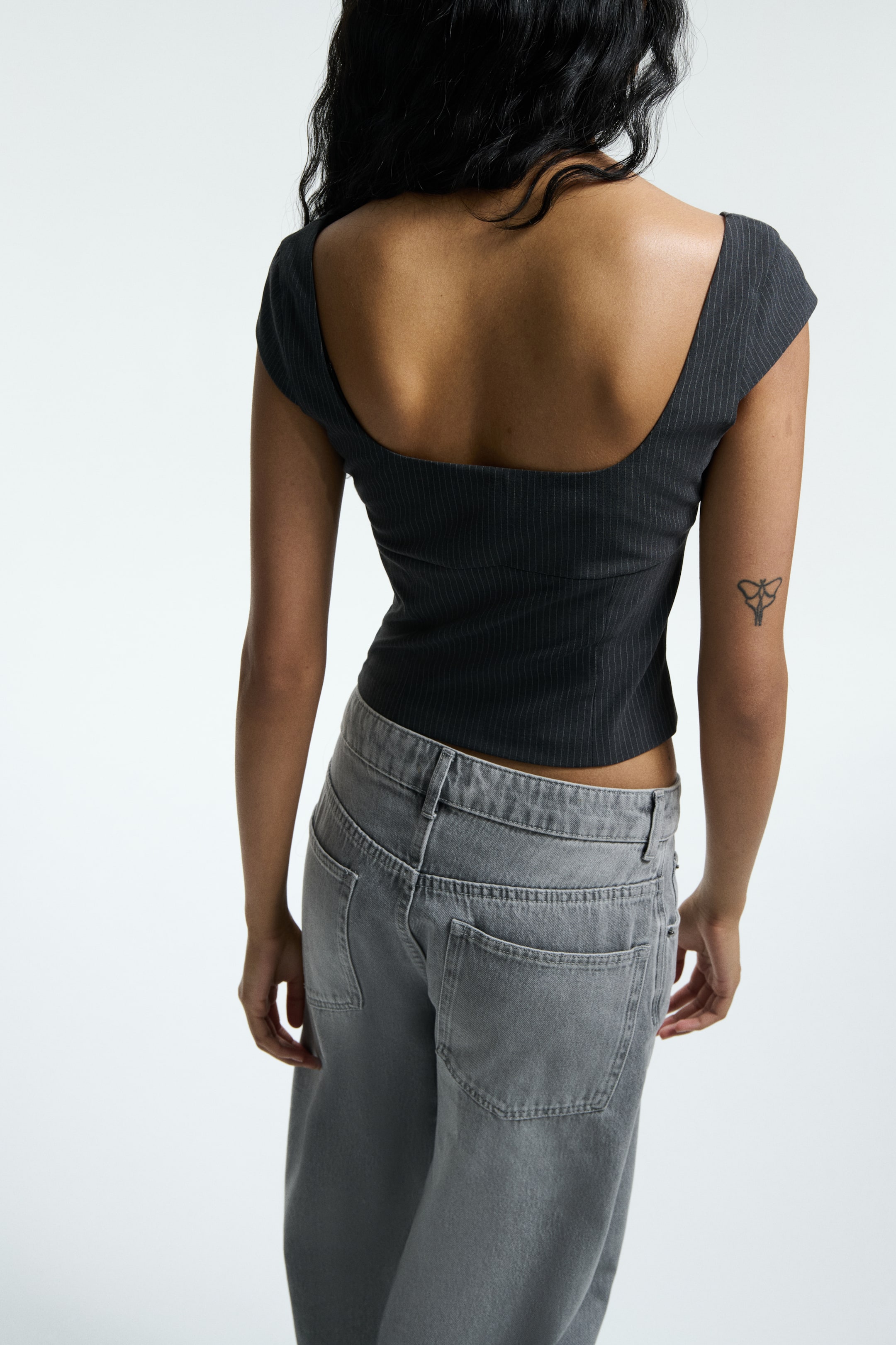 Ver imagen más grande: Un top oscuro a rayas, mostrado desde atrás, presenta una espalda ancha con corte cuadrado y mangas cortas, ajustándose cerca del cuerpo y recortándose en la cintura. Unos jeans holgados de color gris lavado presentan bolsillos traseros visibles, sentándose a media altura por debajo del top.