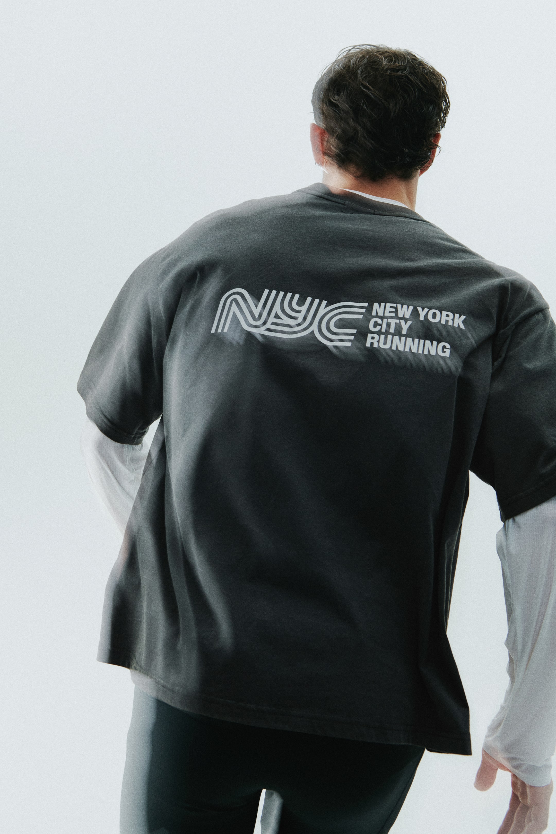 Ampliar la imagen: La modelo lleva puesta una camiseta de manga corta color gris oscuro con un gráfico blanco estilizado de "NYC NEW YORK CITY RUNNING" en la espalda. La camiseta está combinada sobre una prenda blanca de manga larga y con mallas deportivas oscuras.