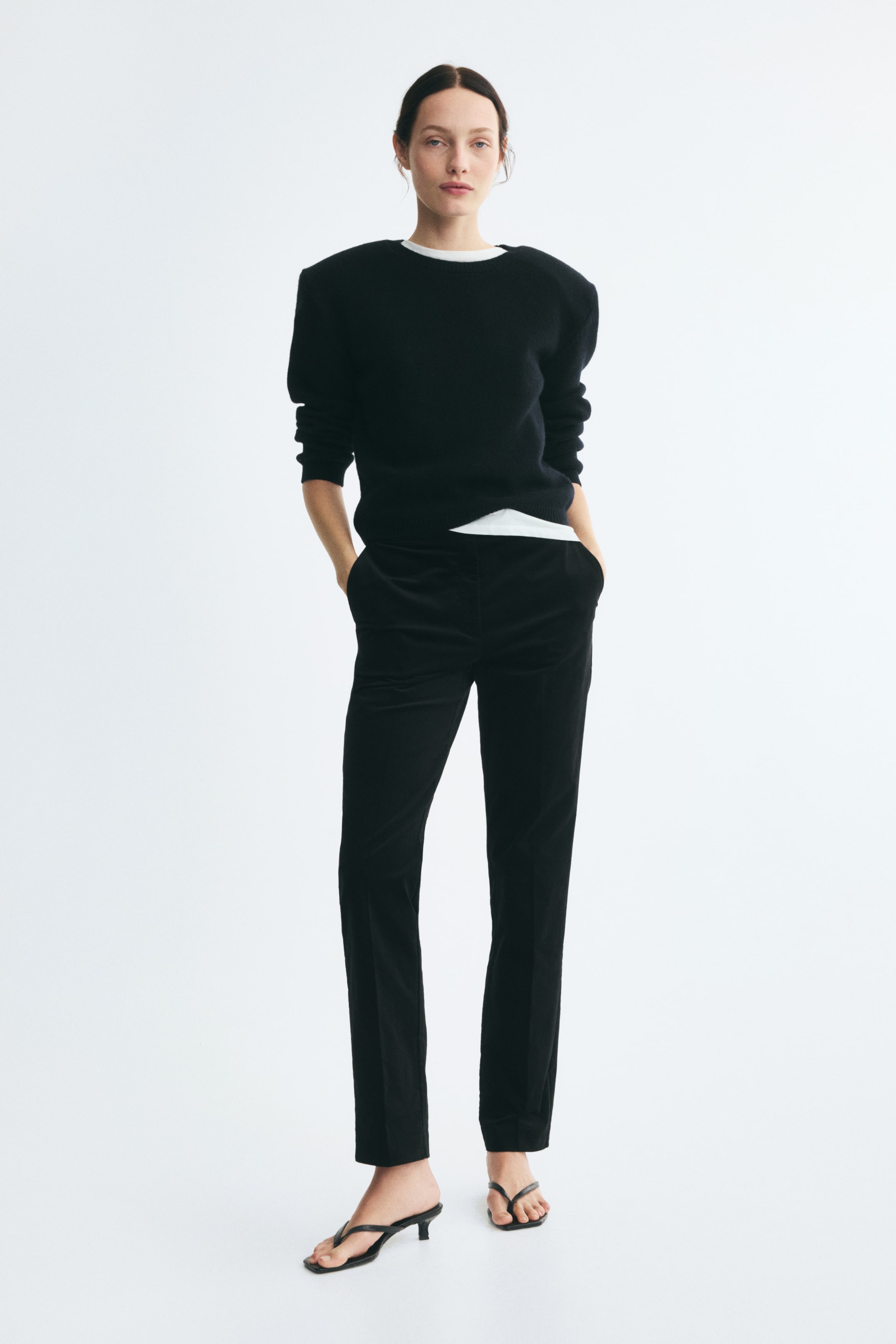 View larger image: Black fitted velvet pants with slim legs and front creases are worn by the model, who stands with her hands resting in the diagonal side pockets. The pants are styled with a black long-sleeve sweater layered over a white top and paired with black heeled sandals.