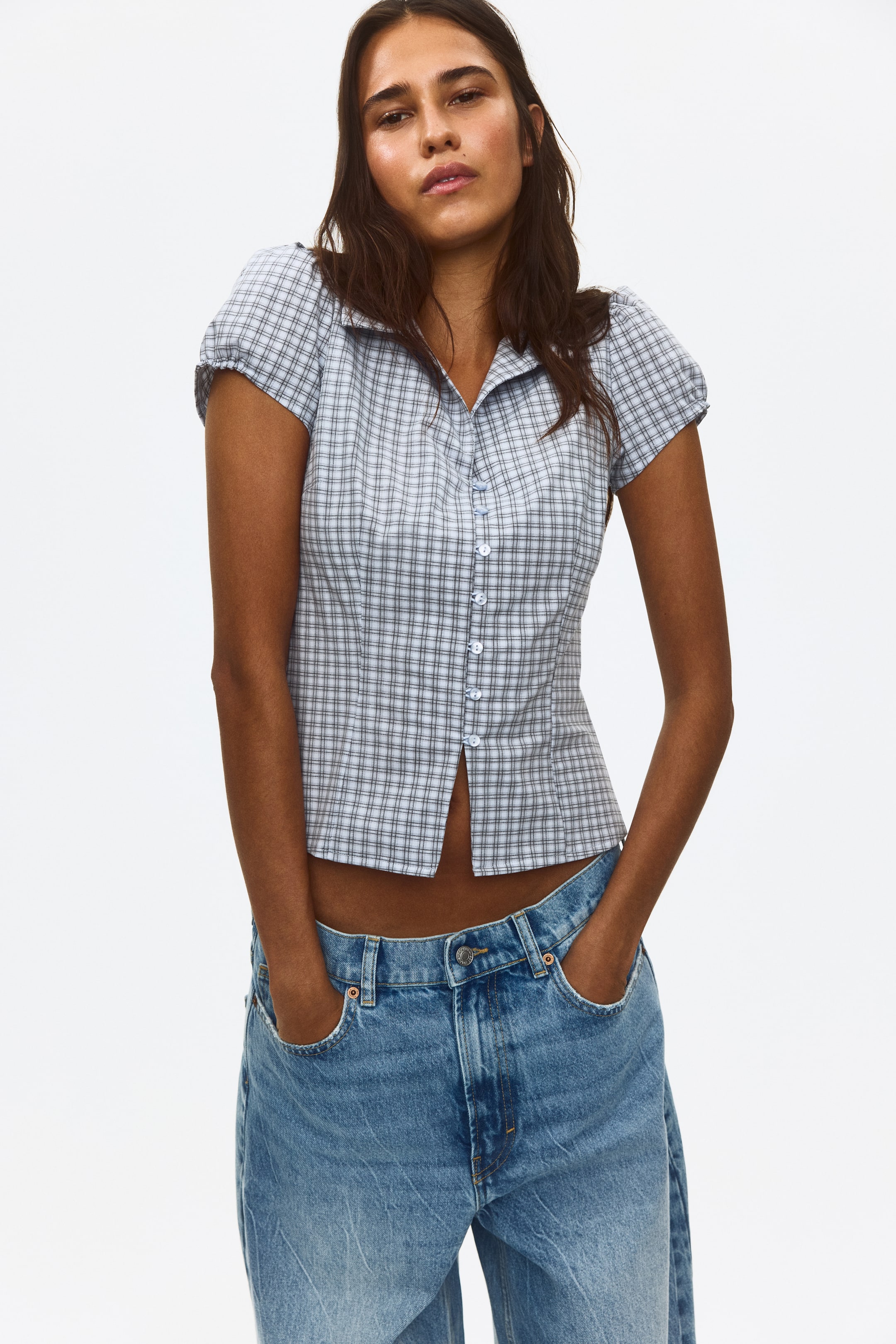 View larger image: Button-front collared top. The model showcases this light blue and white plaid top, featuring a pointed collar, short puffed sleeves, and a cropped hemline. The bottom buttons of the top are undone, revealing the model's midriff above blue jeans.