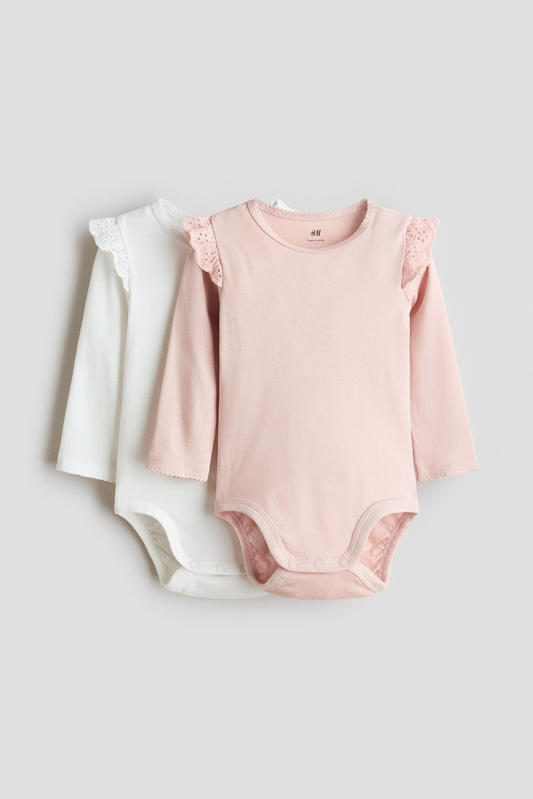 Voir une image plus grande: Deux cache-couches pour bébé sont présentés de face, avec un cache-couche rose pâle à manches longues superposé devant un blanc. Le cache-couche rose présente des volants en dentelle ajourée sur les épaules, des poignets festonnés, un col rond festonné et des fermetures à pression à l'entrejambe. Le cache-couche blanc montre des détails similaires.