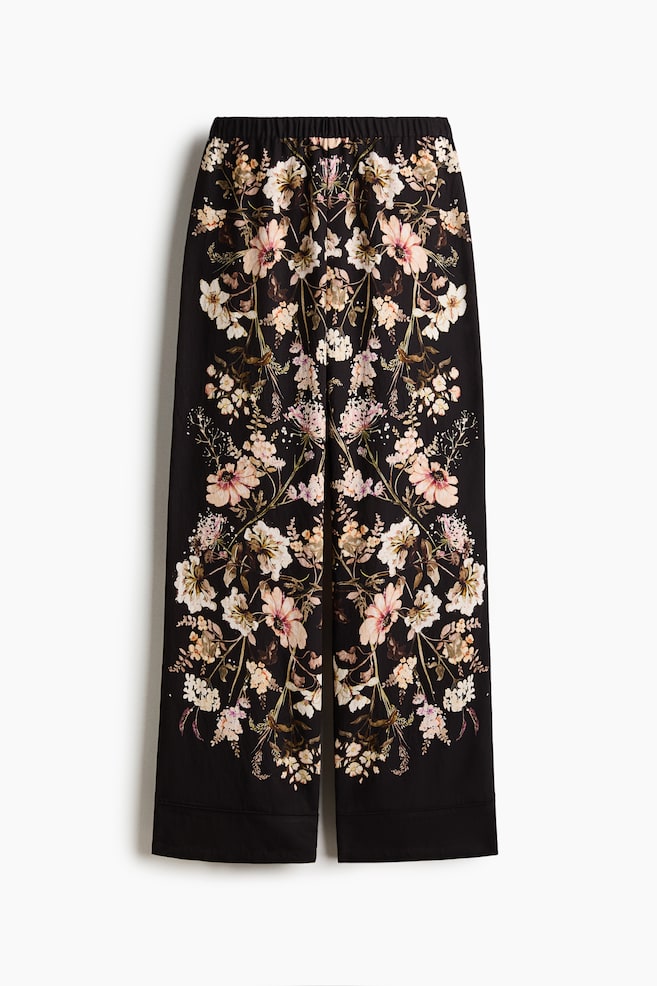 Die Hose weist eine schwarze Grundfarbe mit einem kunstvollen, symmetrischen Blumenmuster aus hellrosa, cremefarbenen und weißen Blüten sowie grünem und braunem Laub auf, das sich von einem elastischen Bund über jedes weite Bein erstreckt. - 1