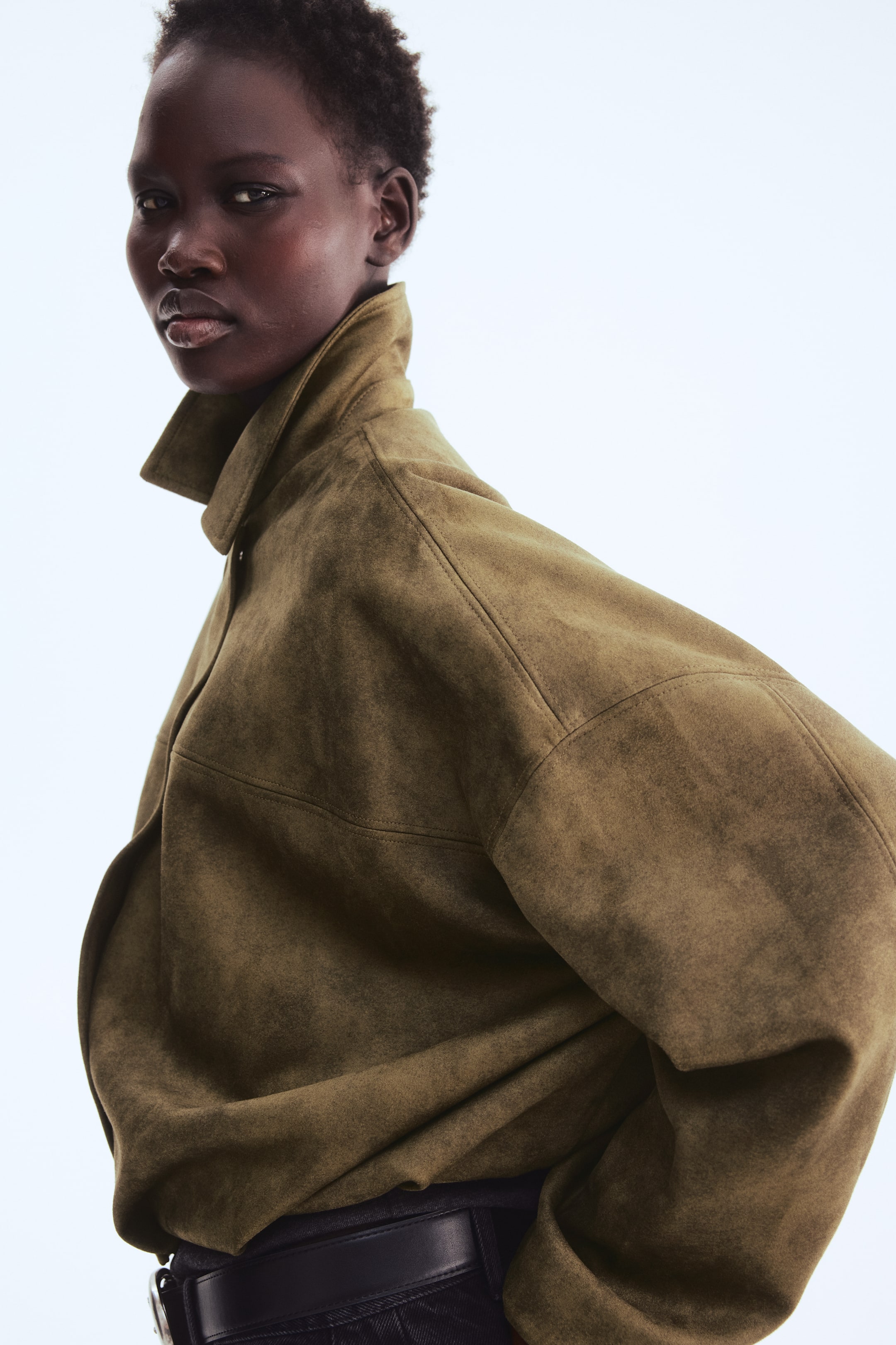 View larger image: The model is wearing an olive green loose-fit jacket made of heavy jersey. The collar stands upright, and a front wind flap is noticeable. A front yoke and low dropped shoulders enhance its puffy silhouette.