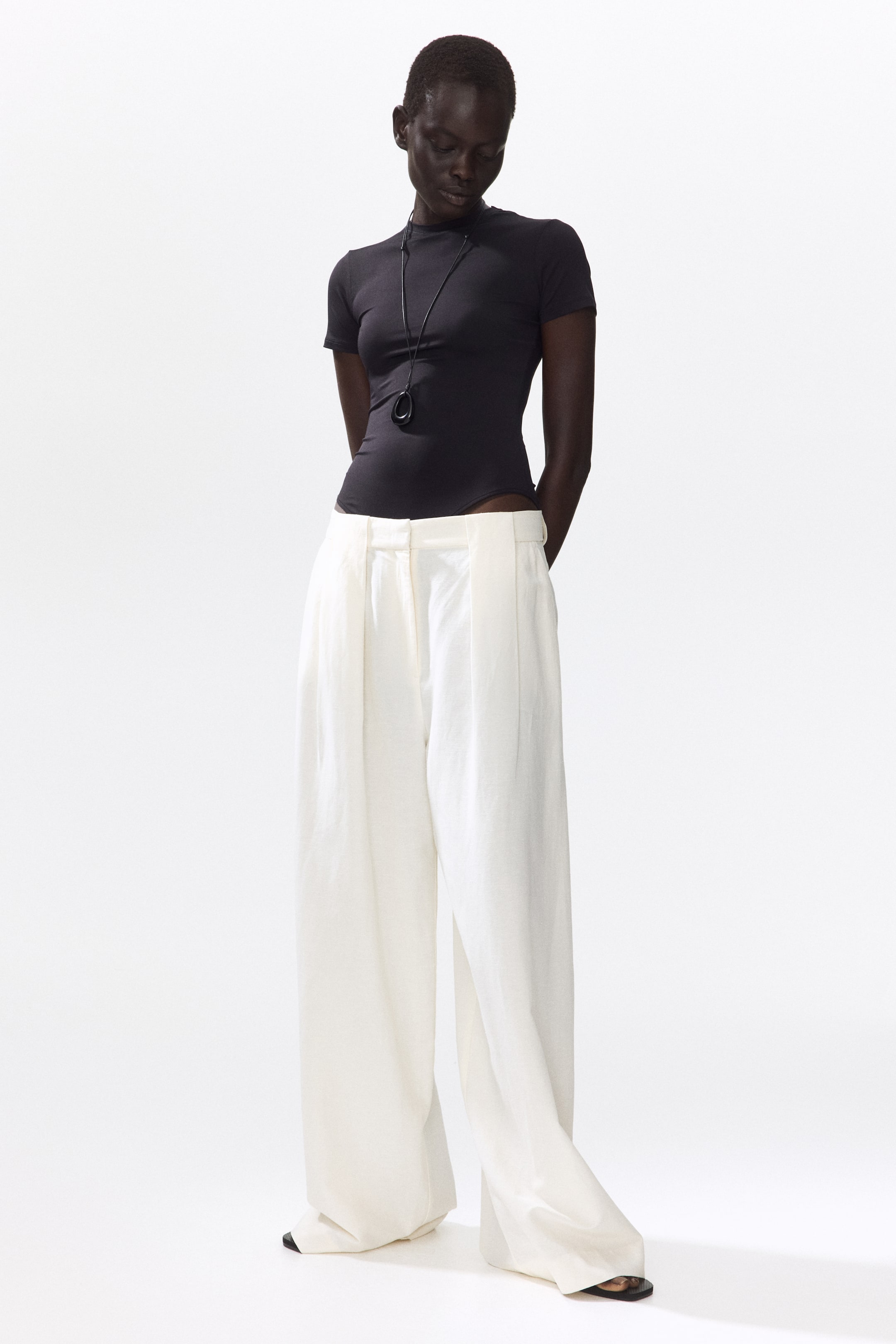 View larger image: A black short-sleeve mock-neck bodysuit fits snugly, paired with a black abstract pendant necklace resting on it. It's tucked into off-white, high-waisted, wide-leg pleated pants that flow to the floor, partially obscuring black sandals.