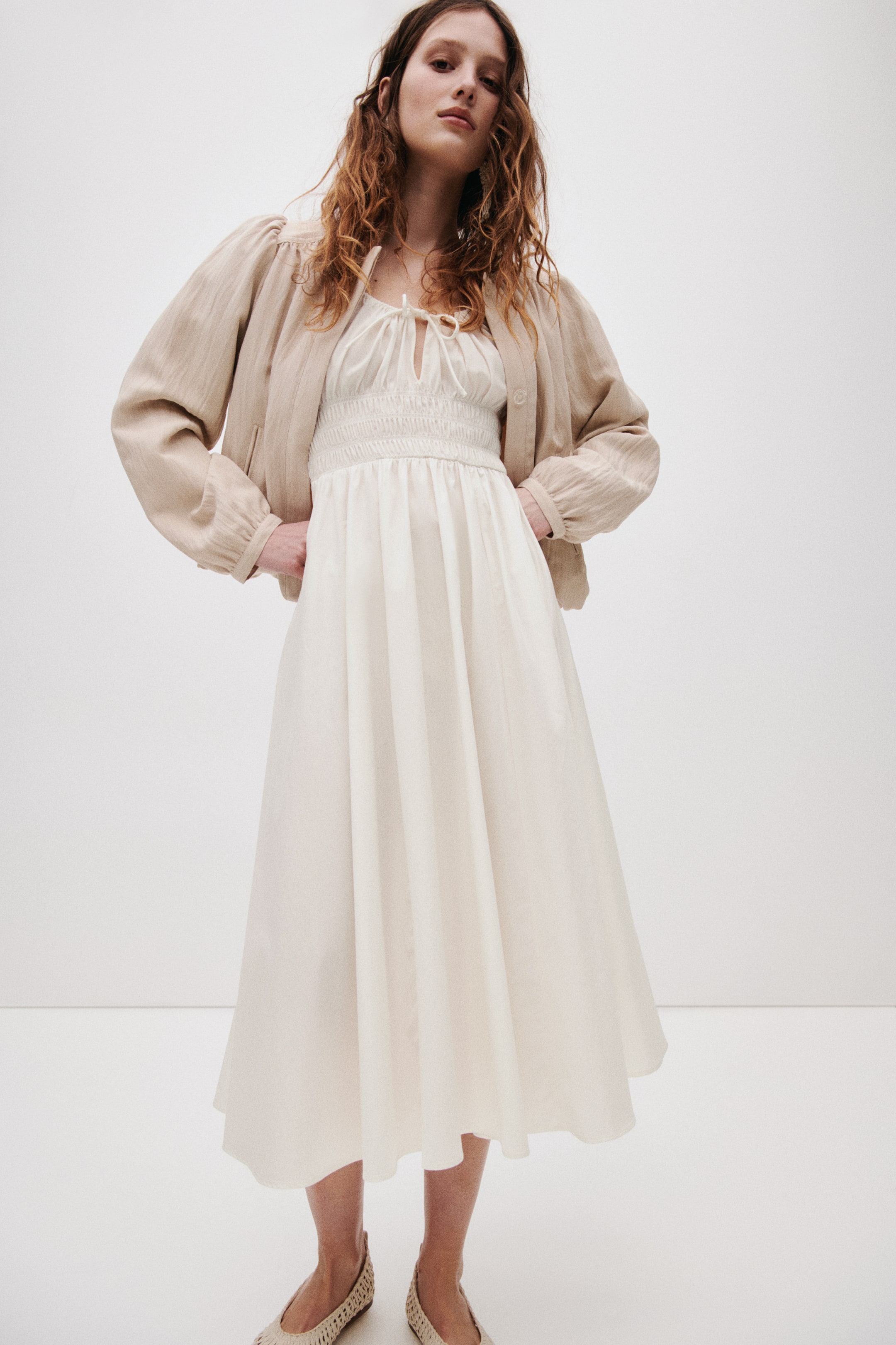 View larger image: A light beige bomber jacket layers over a cream midi dress. The jacket features long puffed sleeves with gathered cuffs, while the dress showcases a smocked bodice with a tie-front V-neckline and a flowing A-line skirt.