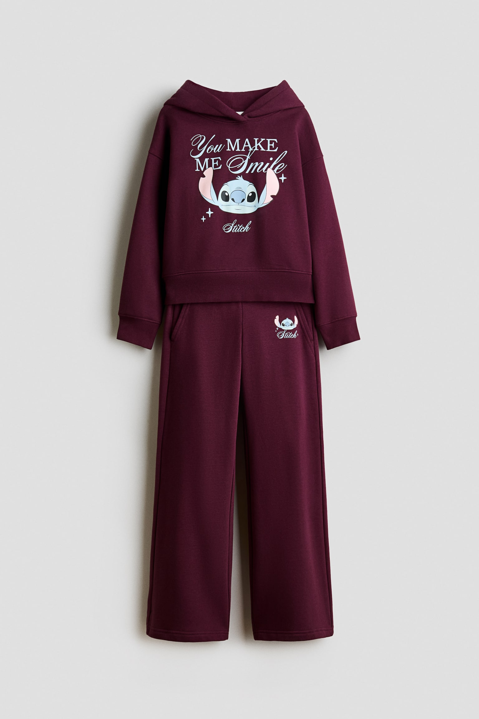 This front-facing view shows a dark purple fleece hoodie and track pants set. The hoodie features a wrap-front hood, dropped shoulders, ribbed trims, and a central Stitch graphic with the text "You MAKE ME Smile," while the matching straight-leg track pants feature diagonal pockets and a small Stitch print on the hip.