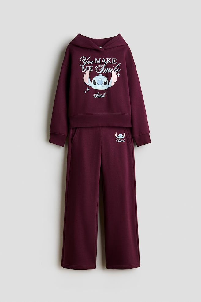 Questa vista frontale mostra un completo composto da hoodie in fleece viola scuro e pantaloni sportivi. La hoodie presenta un cappuccio con davanti avvolgente, spalle scese, finiture a costine e una grafica centrale di Stitch con la scritta "You MAKE ME Smile", mentre i pantaloni sportivi abbinati a gamba dritta mostrano tasche diagonali e una piccola stampa di Stitch sul fianco. - 1