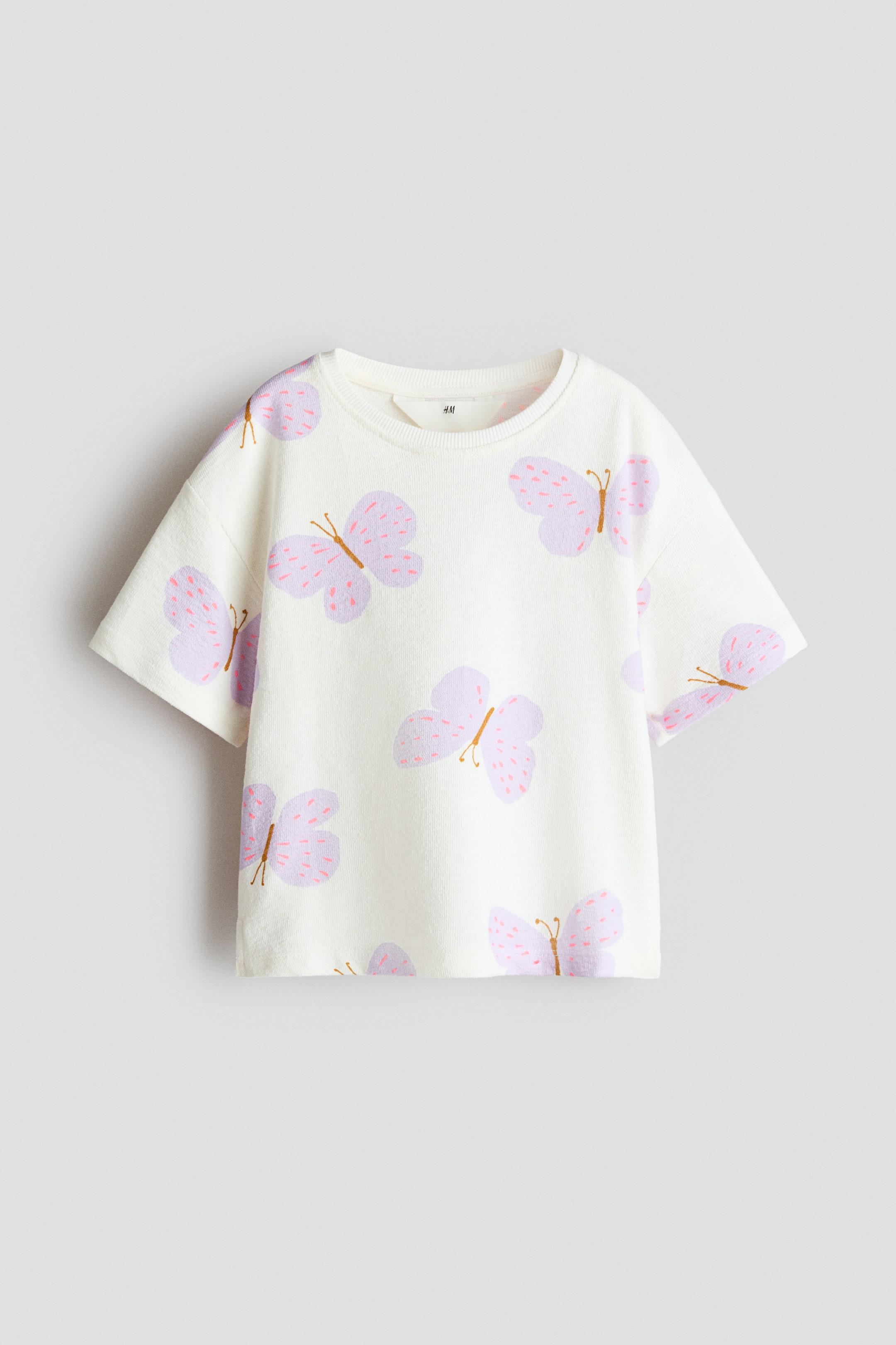 Größeres Bild ansehen: Ein cremefarbenes, strukturiertes Jersey-T-Shirt zeigt seine Vorderansicht und verfügt über einen runden, gerippten Halsausschnitt, überschnittene Schultern, kurze Ärmel und ein durchgehendes Muster aus hellvioletten Schmetterlingen mit rosa Sprenkeln.