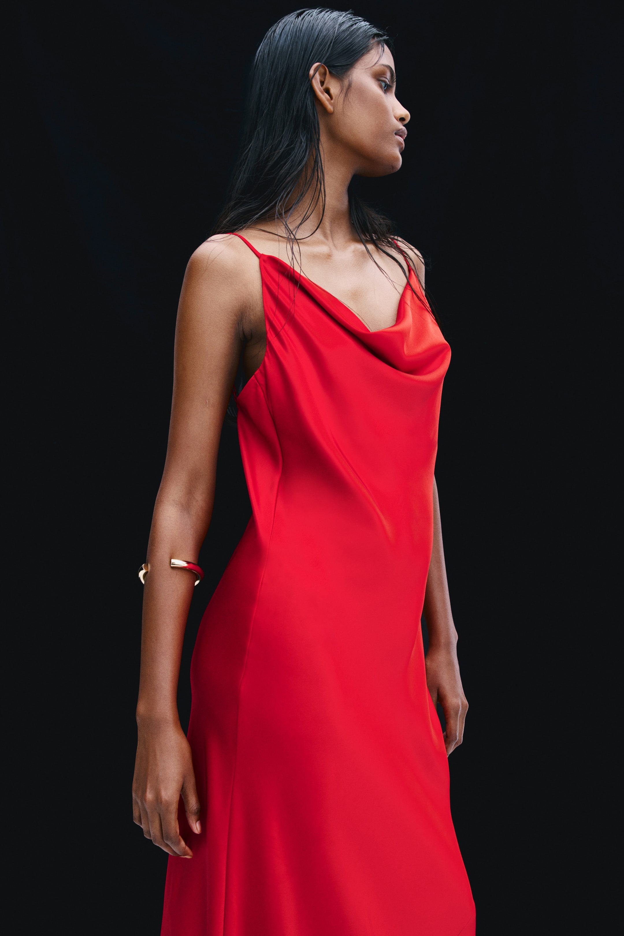 View larger image: The model is wearing a red satin dress featuring a draped cowl neckline and thin spaghetti straps. The softly draped, fitted dress is styled with a gold-coloured cuff bracelet on the model's forearm.
