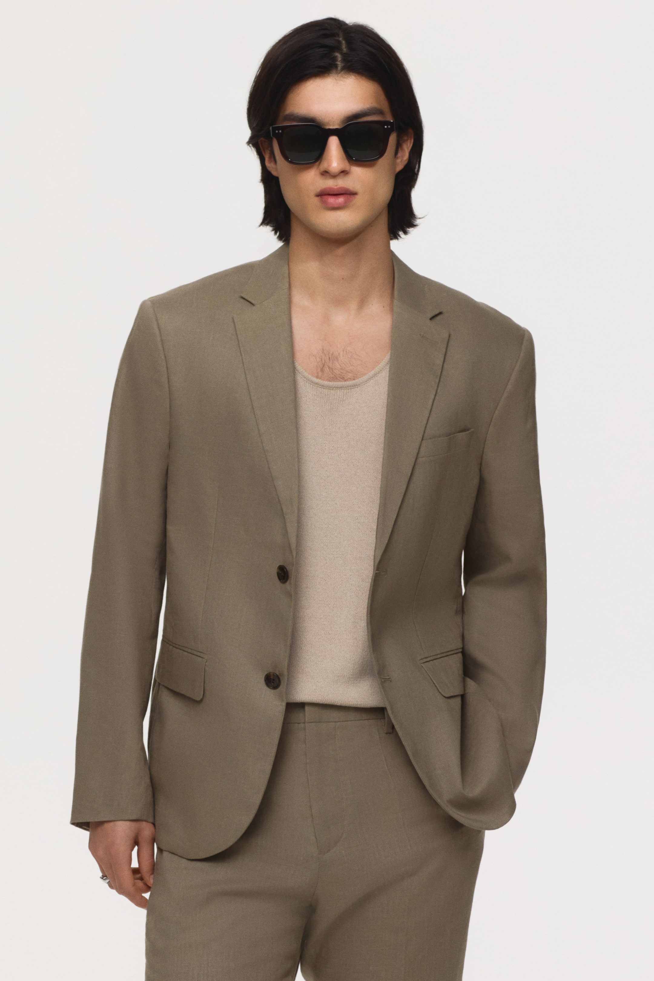 View larger image: A taupe single-breasted suit crafted from a textured fabric is showcased front-facing, featuring a two-button closure, notch lapels, and flap pockets, paired with matching trousers. It is styled over a light beige ribbed tank top.