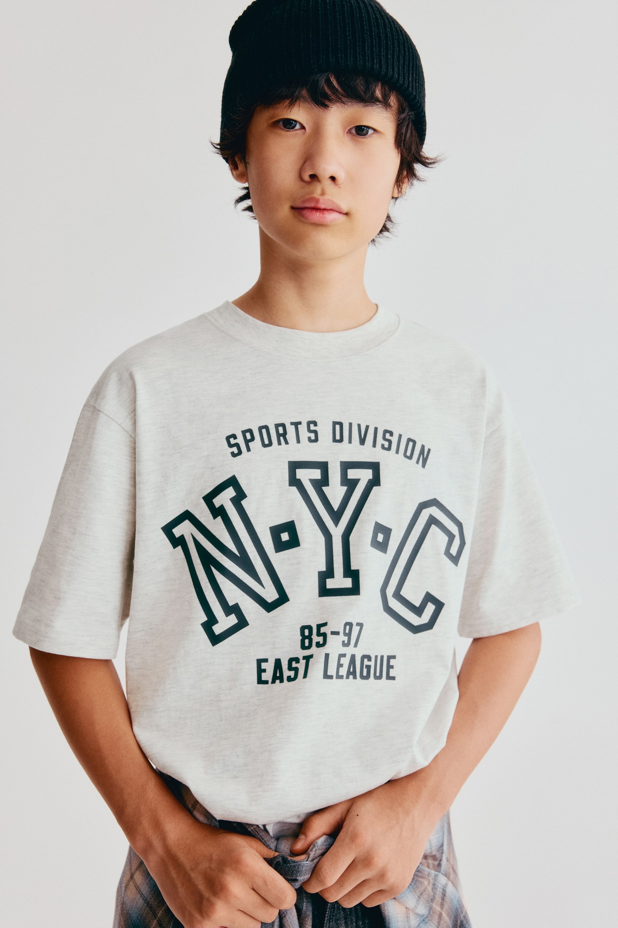 拡大画像を表示: フロントビューで着用された、ゆったりとしたフィット感のコットンジャージーTシャツ。リラックス感のあるシルエット、ドロップショルダー、リブ仕様のクルーネックラインが際立っています。ライトグレーのTシャツの前面には、ダークカラーのカレッジ風グラフィックプリントが施されており、「N-Y-C」の文字と「SPORTS DIVISION」「EAST LEAGUE」のテキストがデザインされています。