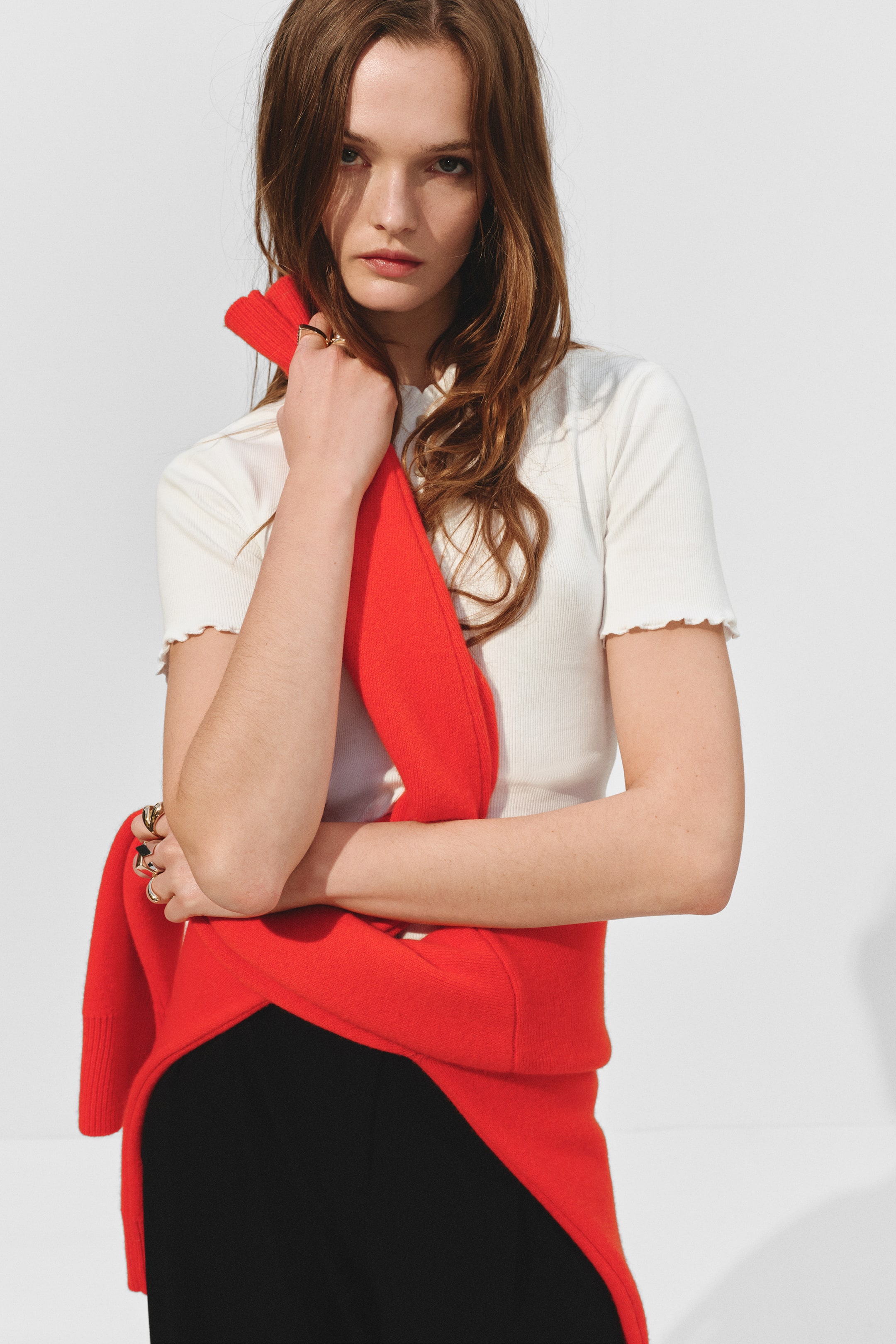 查看大圖: 一件紅色針織毛衣從左肩到右臀斜披在身上,袖子環繞著腰部,搭配在白色羅紋上衣和黑色下裝之上。