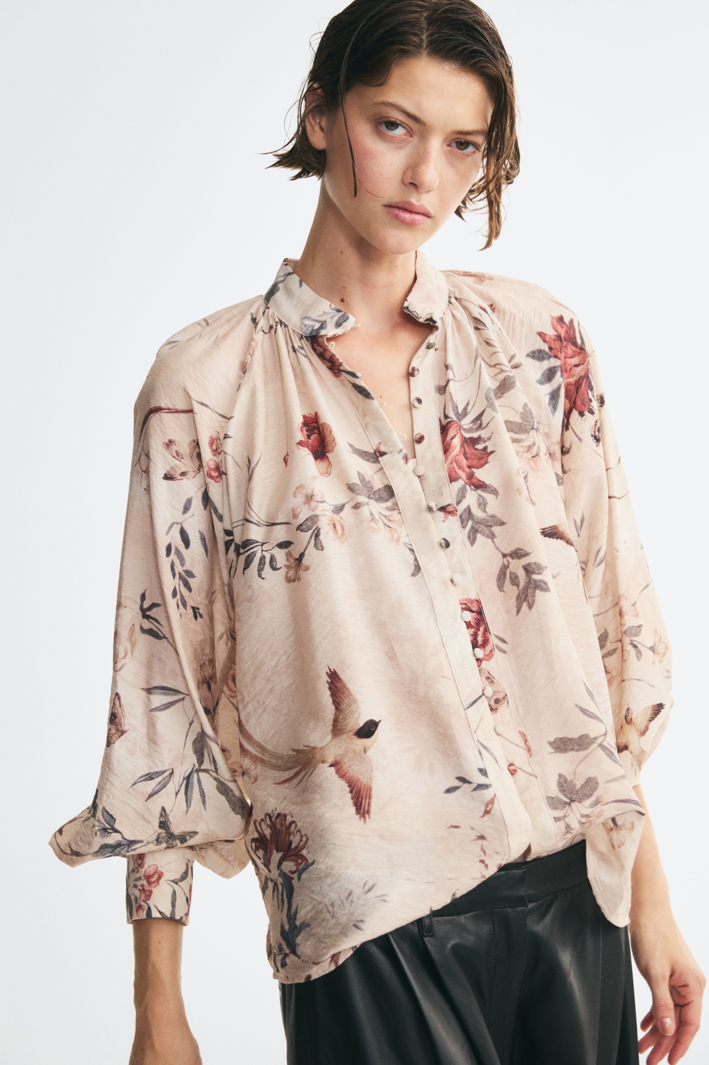 An oversized beige blouse is shown from the front, showcasing a botanical floral pattern in muted red and dark grey tones. It features a gathered stand collar, a front placket with covered buttons, and long balloon sleeves ending in wide button cuffs.