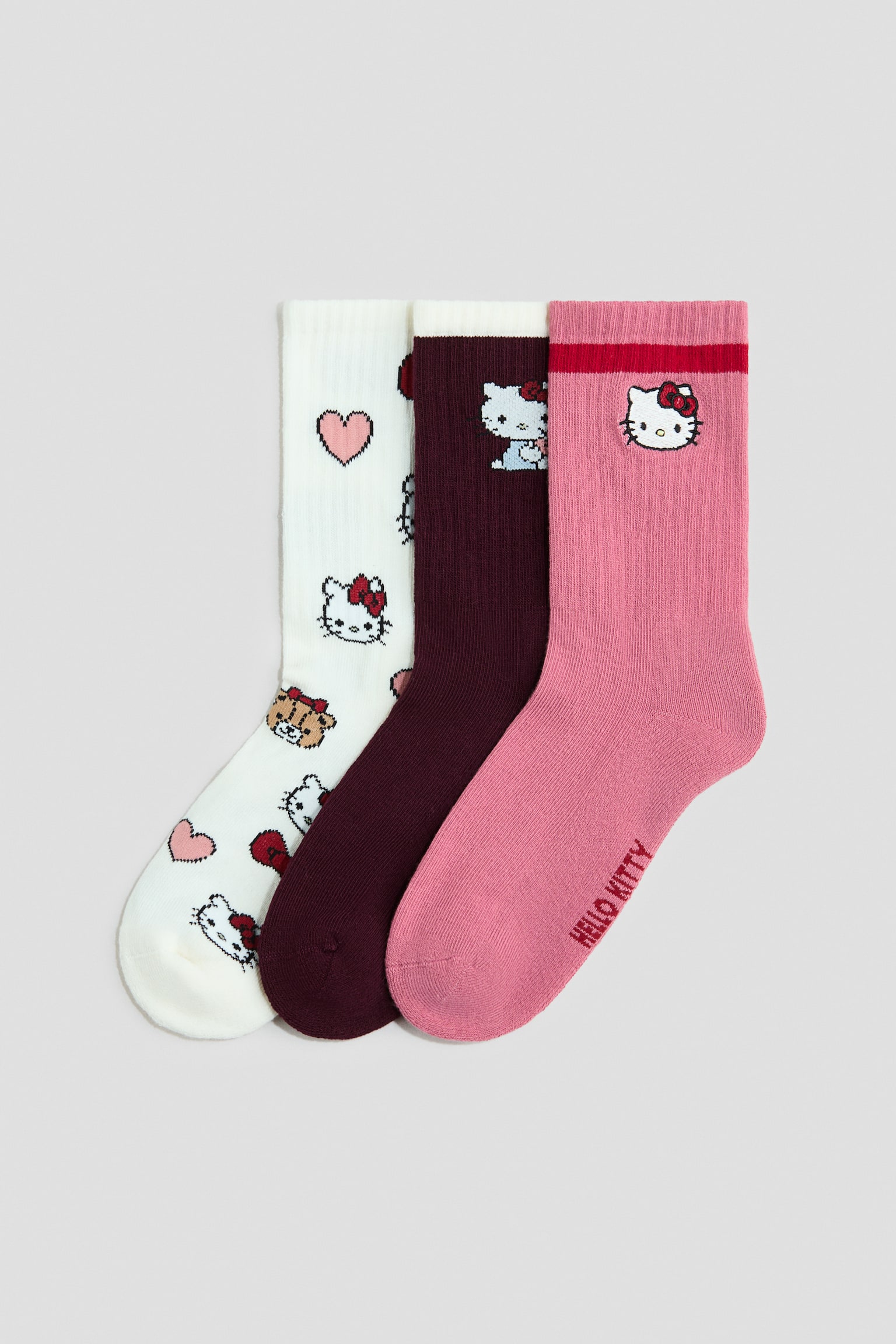 Trois chaussettes en maille fine sont posées côte à côte de profil, avec des tiges côtelées et des bords élastiqués. L’ensemble comprend une chaussette blanc cassé ornée d’un motif intégral de cœurs et de visages de personnages, une chaussette bordeaux foncé avec un motif de personnage unique près de la bordure, et une chaussette rose présentant une rayure rouge, un visage de personnage et du texte le long de la semelle.