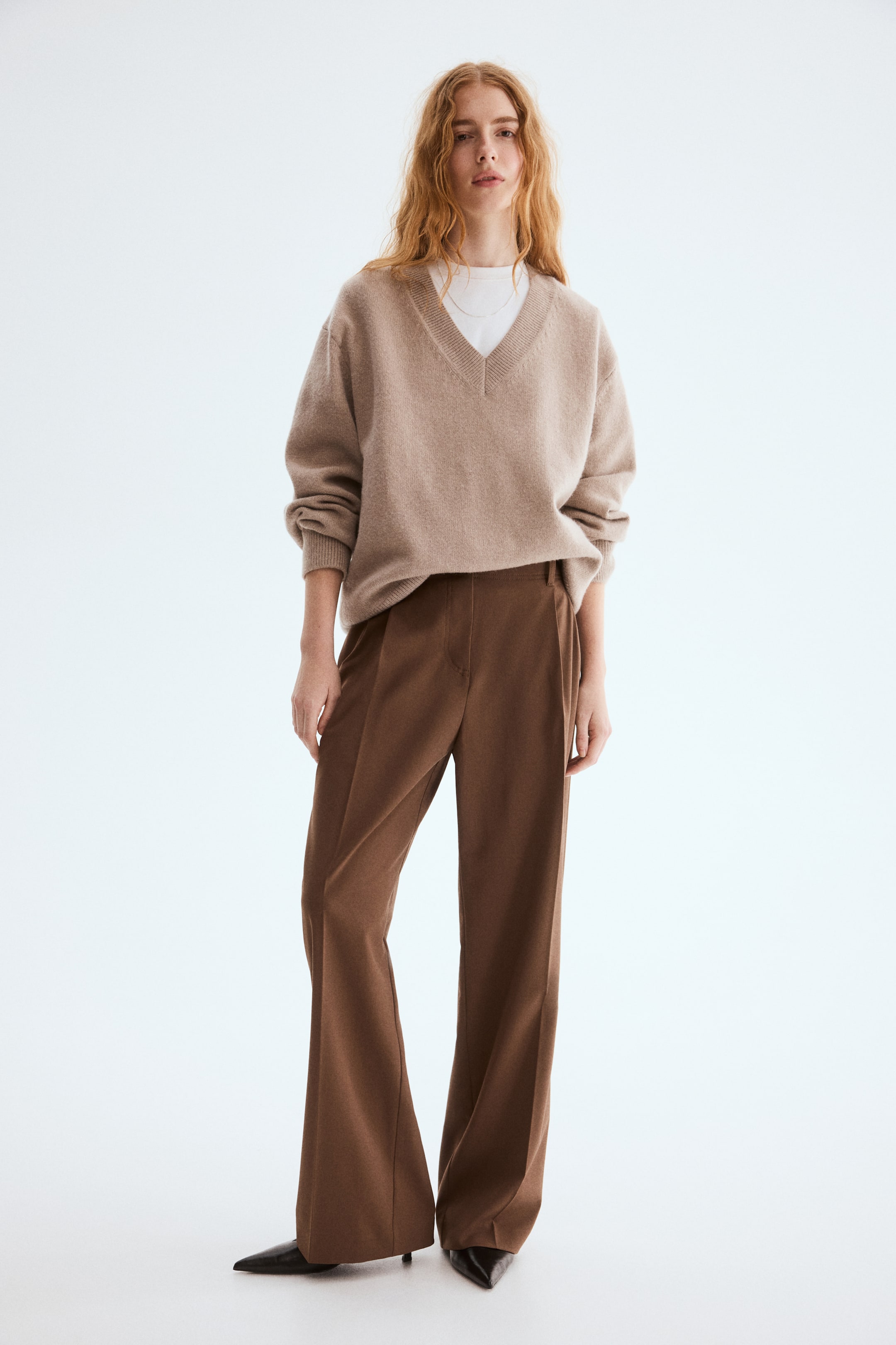 View larger image: A light brown v-neck sweater, featuring a relaxed fit and ribbed trim at the neckline, cuffs, and hem, is being worn over a white crew-neck t-shirt. Brown wide-leg trousers, featuring front pleats, are being styled with the sweater.