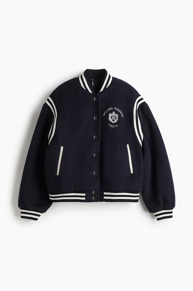 Eine dunkelblaue Collegejacke wird von vorne präsentiert, mit einem gerippten schwarz-weiß gestreiften Stehkragen, Bündchen und Saum. Sie zeigt einen Druckknopfverschluss vorne, weiße Streifendetails an den langen Ärmeln, zwei weiß eingefasste vordere Leistentaschen und ein weißes gesticktes Wappen auf der Brust. - 1