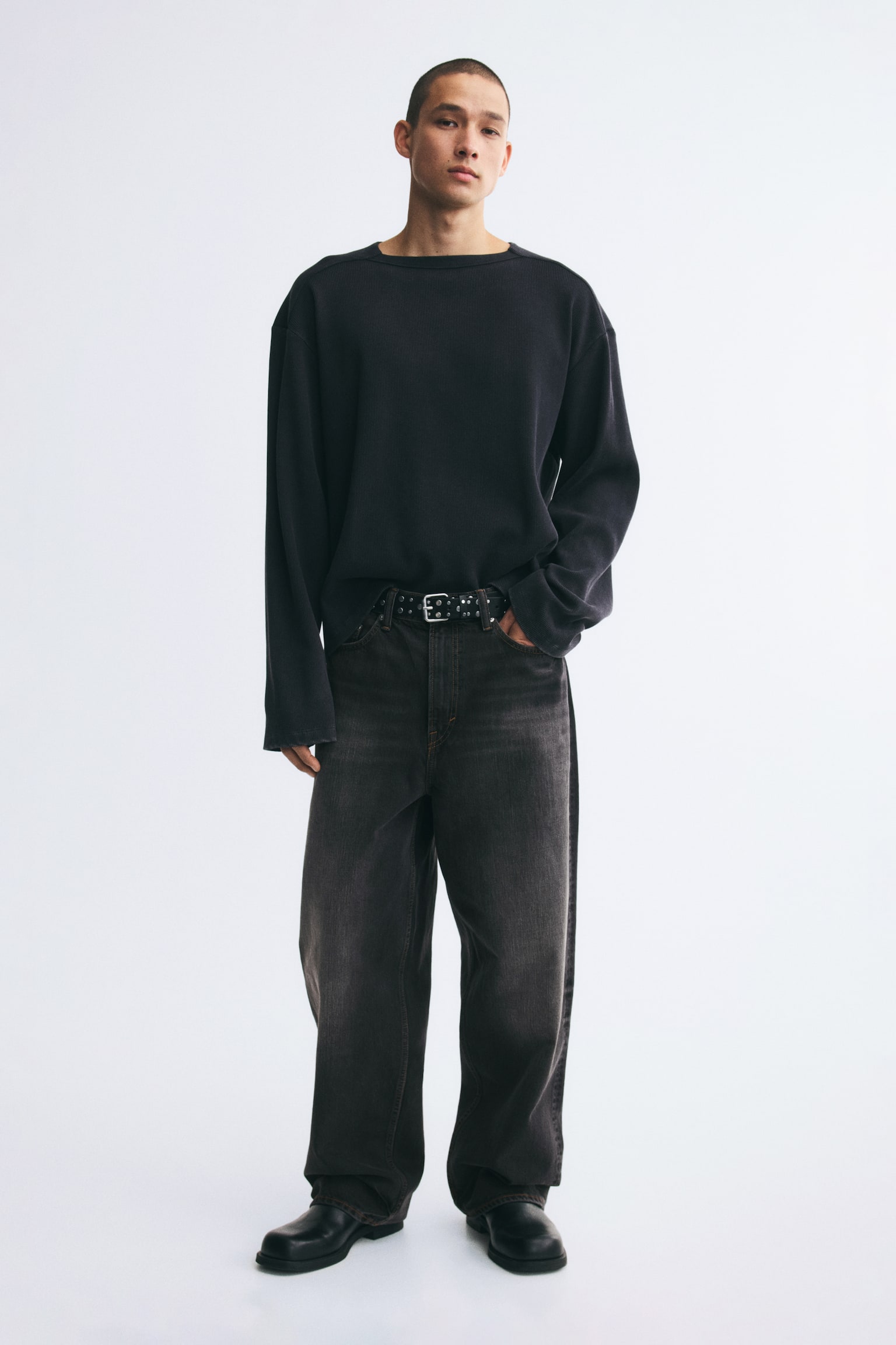 The model is wearing faded dark grey, baggy-fit denim jeans, featuring a relaxed, rounded-leg silhouette that stacks at the ankles. The jeans are styled with a black studded belt at the waist, a black long-sleeve top, and black boots. - 1