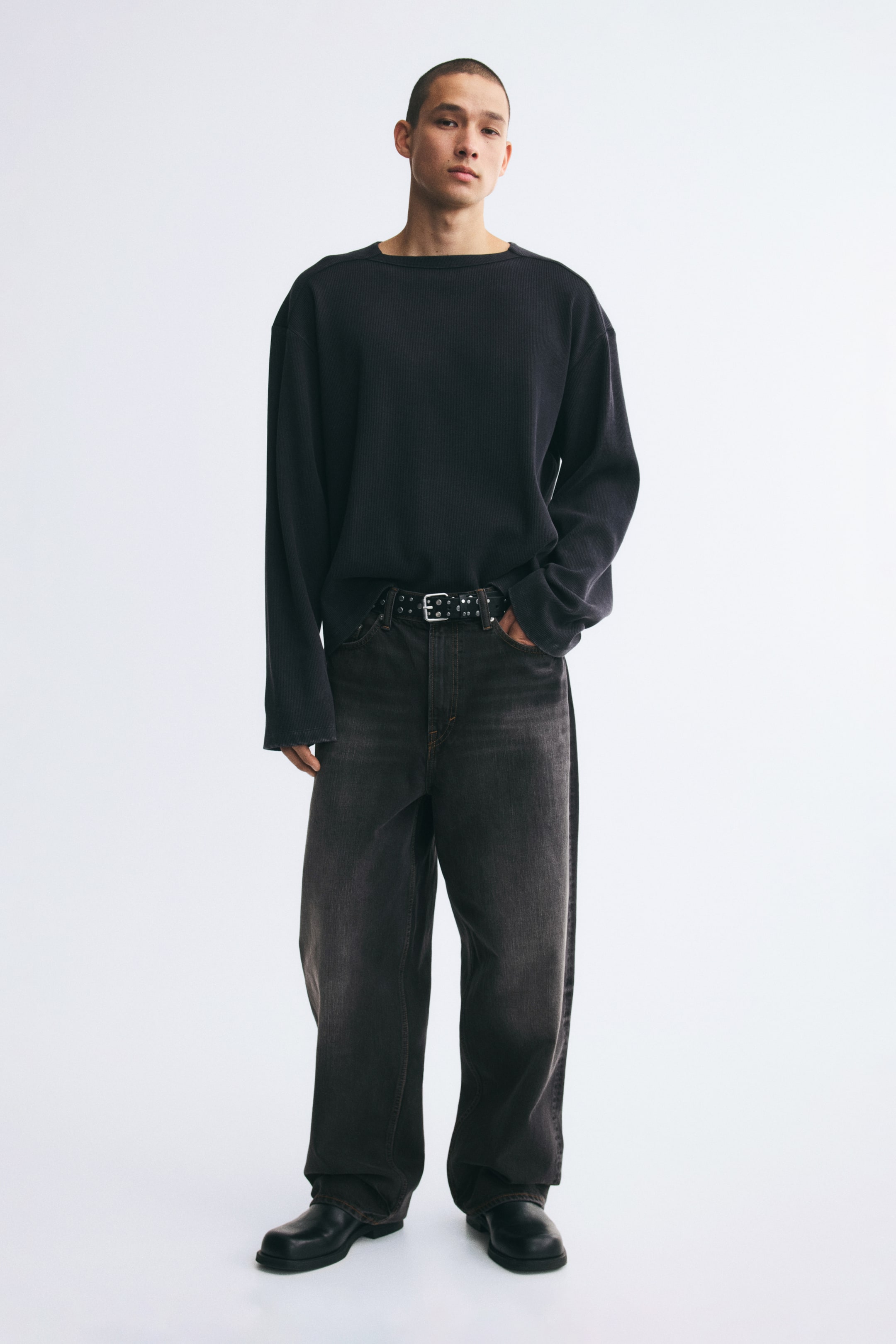 Xem ảnh lớn hơn: Quần jeans denim màu xám đậm bạc màu, phom baggy, đang được người mẫu mặc, nổi bật với dáng ống rộng, bo tròn và xếp nếp ở mắt cá chân. Quần được phối cùng thắt lưng đen đính đinh tán ở eo, áo tay dài màu đen và bốt đen.